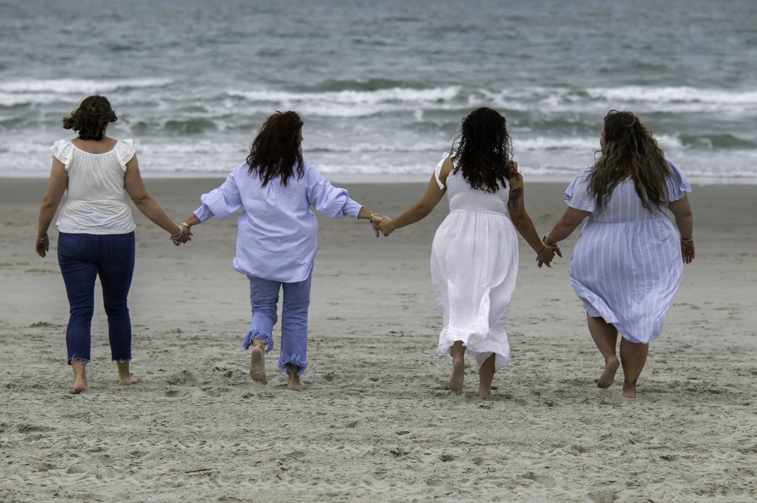 Four women holding hands and walking barefoot on the beach, facing the ocean.