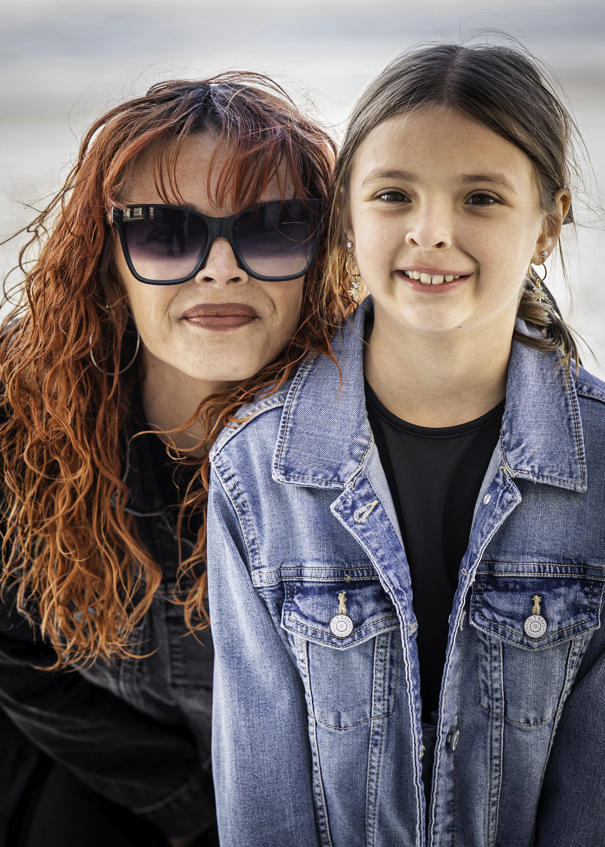 A woman with red hair wearing large sunglasses and a black top, standing next to a girl with brown hair in pigtails, wearing a denim jacket and smiling at the camera. They are outdoors near the beach.