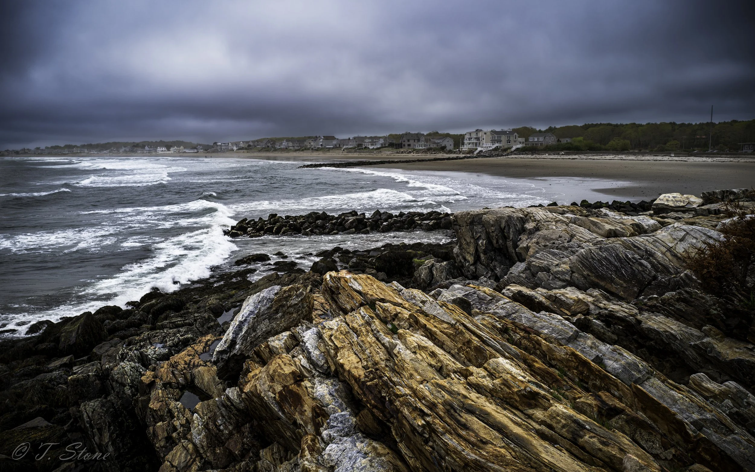 Rocky shoreline with waves crashing against the rocks, dark overcast sky, and houses along the distant beach.