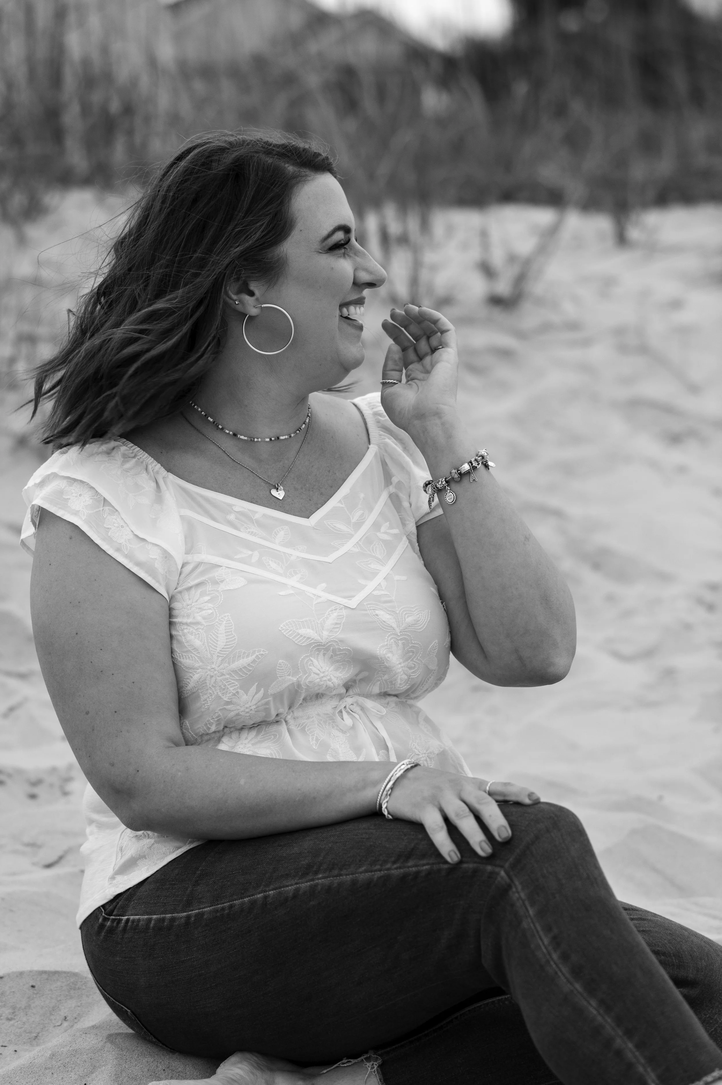 A woman with long hair sitting on the sand and smiling. She is wearing hoop earrings, a necklace, and a bracelet, and has her hand near her face. The background shows a blurred outdoor scene with trees.