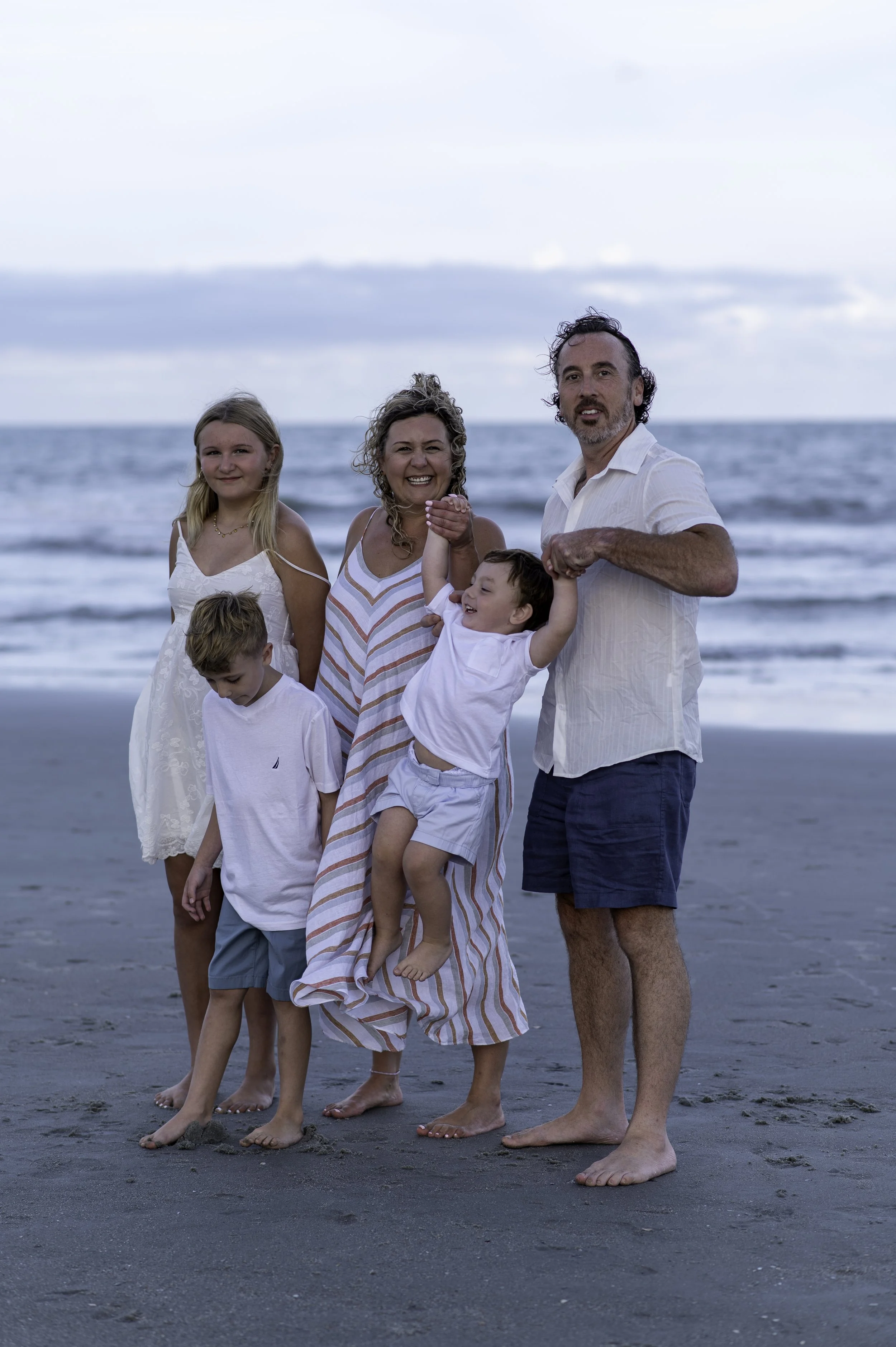 A family of six standing barefoot on the beach with sand, with the ocean and cloudy sky in the background. The group includes three children and three adults, all smiling and dressed in light summer clothes.