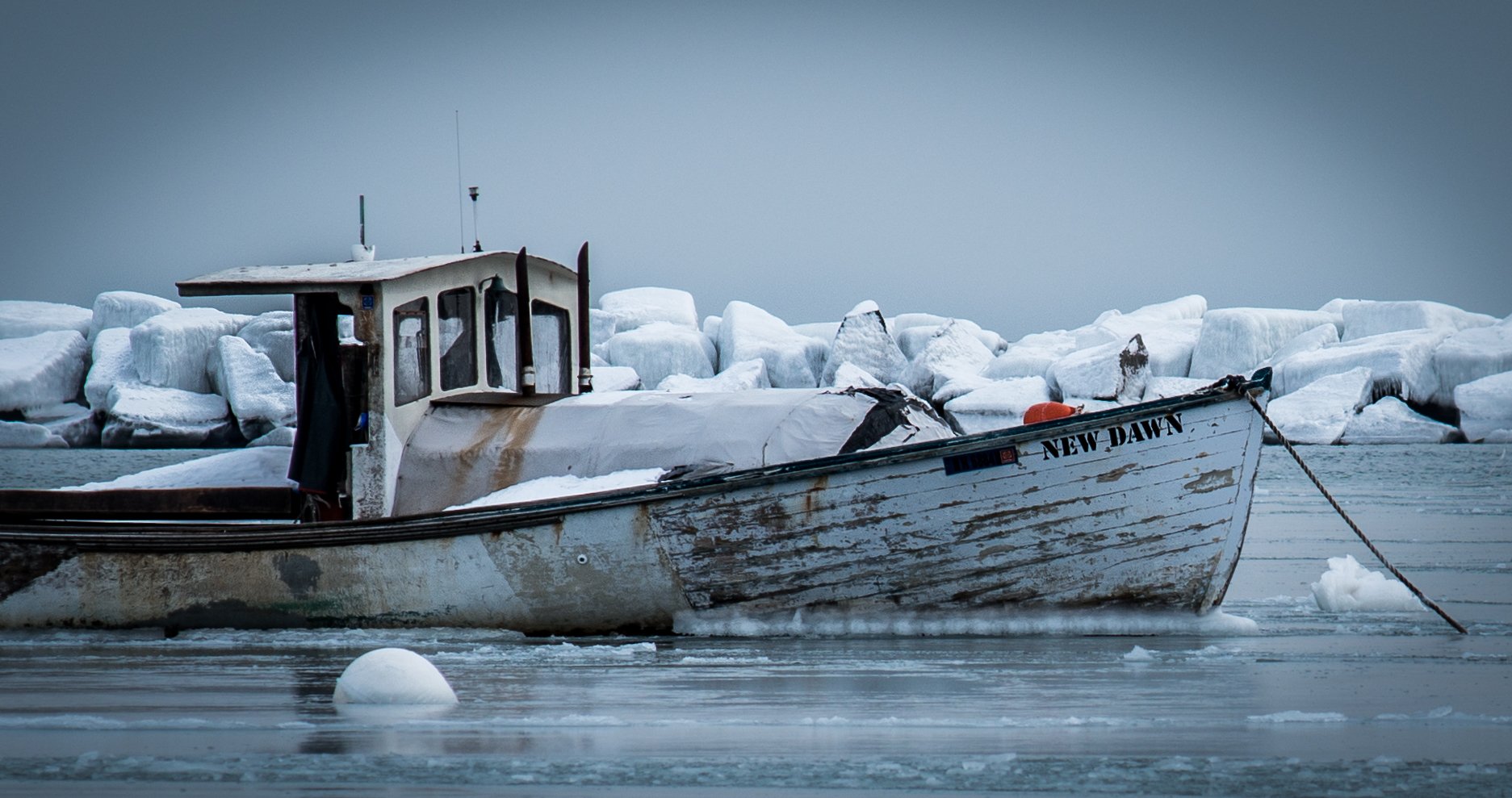 A weathered boat named New Dawn moored in icy water with large ice chunks and a cloudy sky in the background.