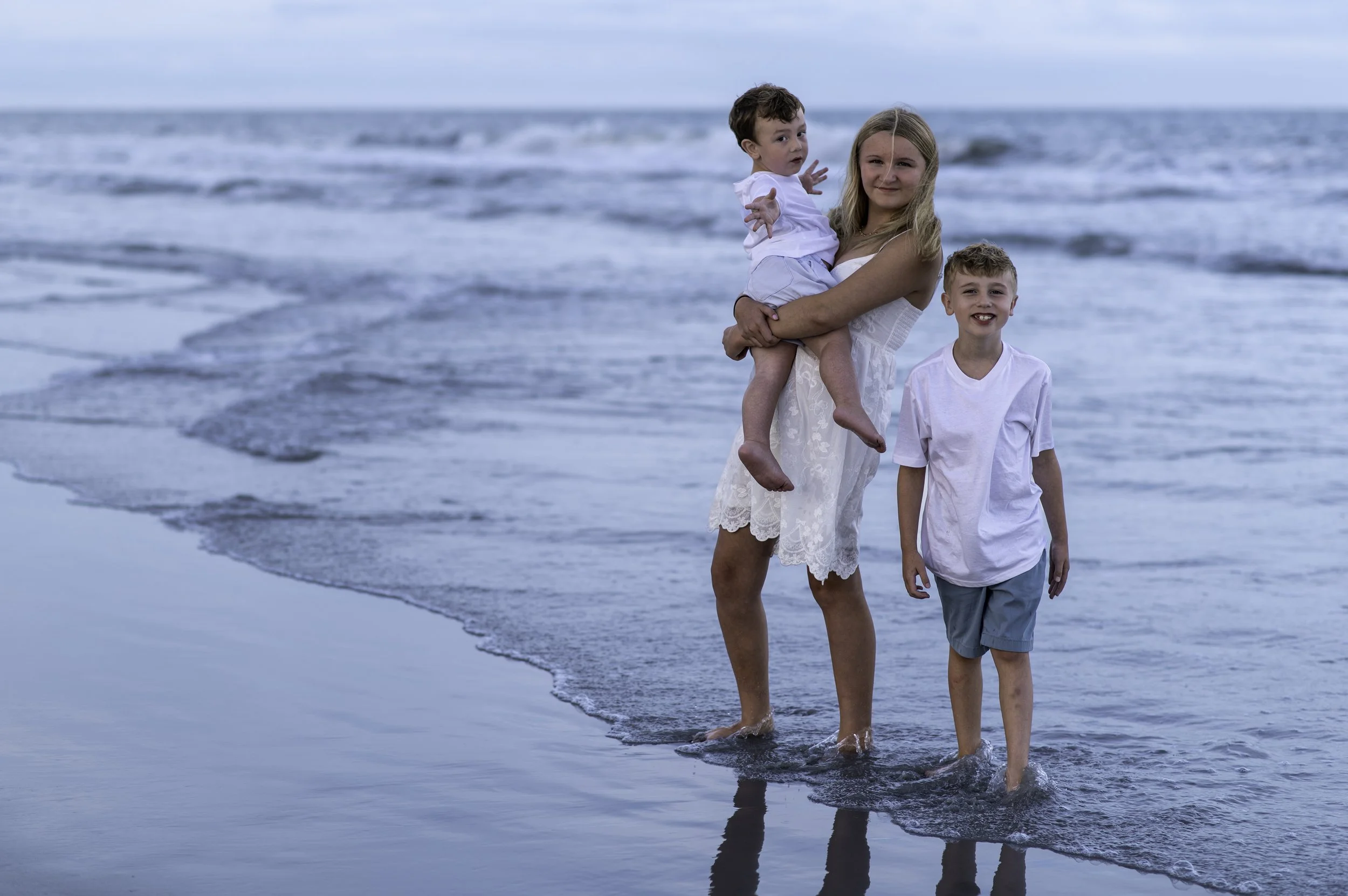 A woman and two children enjoying a walk along the beach with waves in the background.