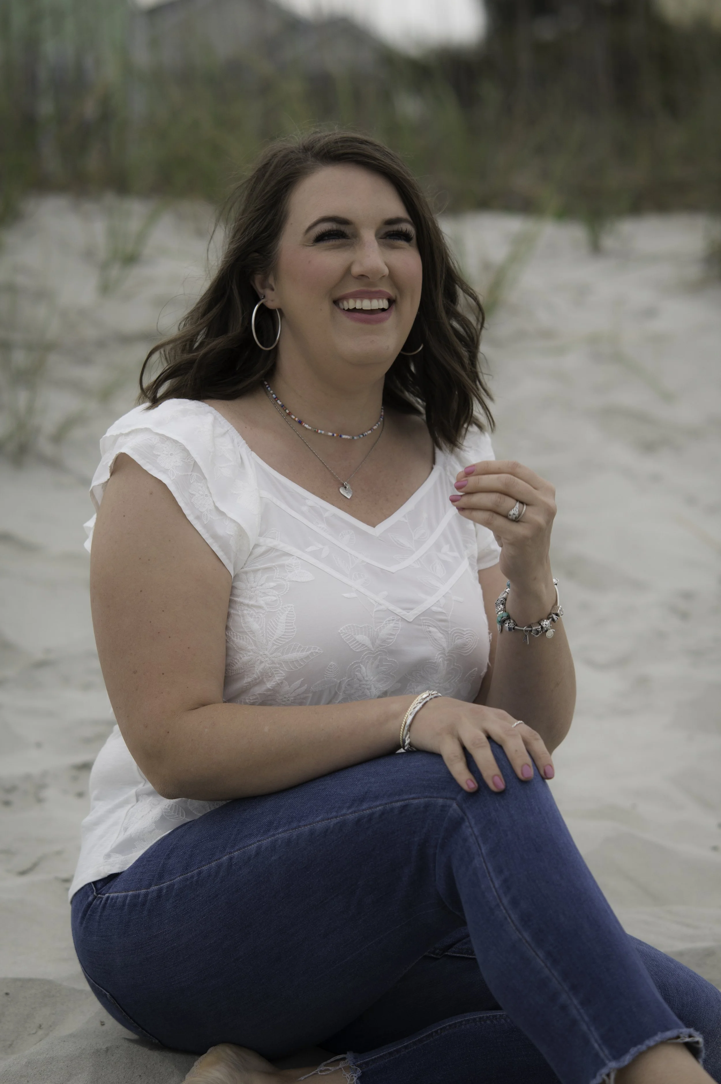 A woman sitting on the sand at the beach, smiling and looking away, wearing a white top, blue jeans, jewelry, and hoop earrings.