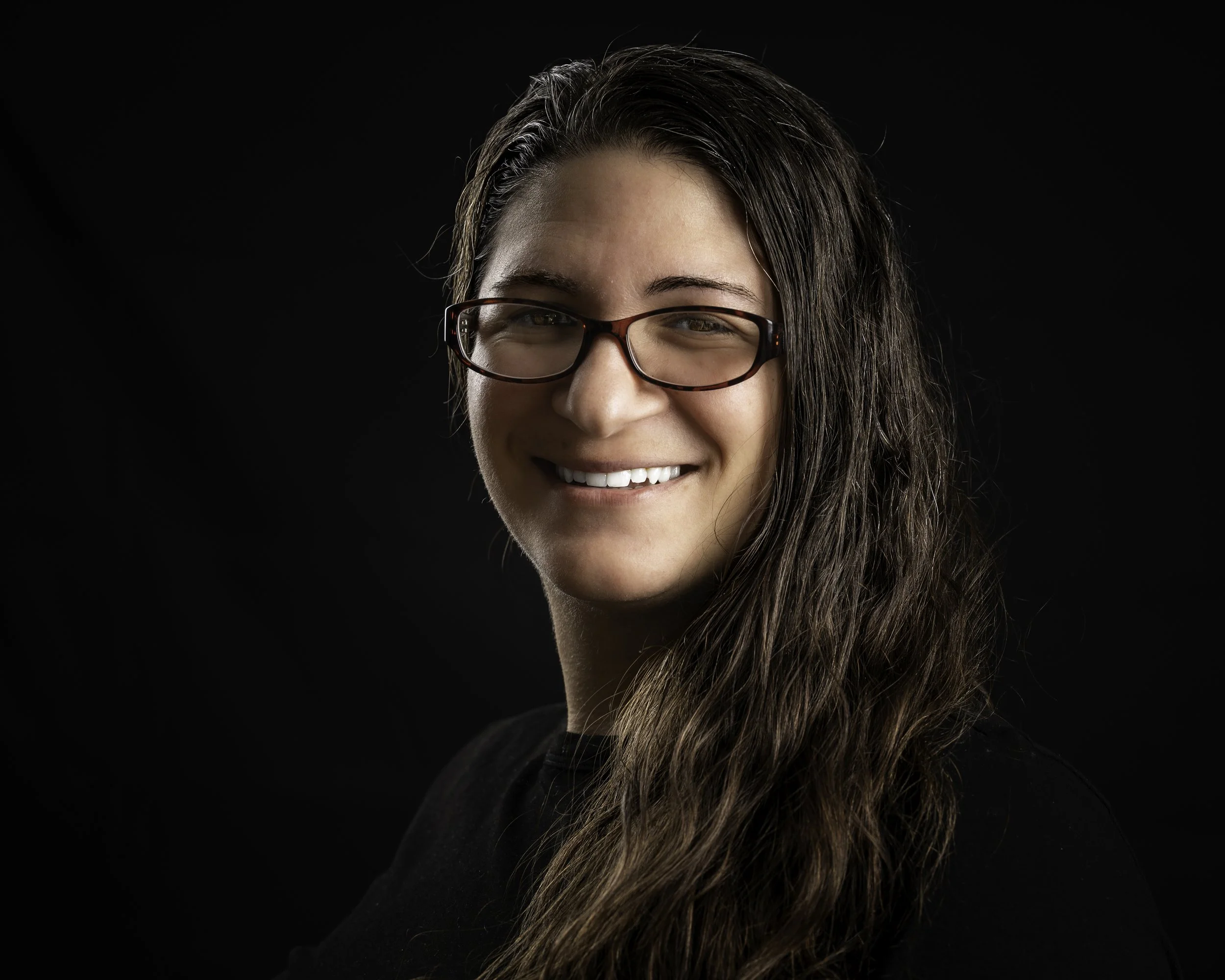 Close-up portrait of a young woman with long wavy hair, wearing glasses, smiling against a black background.