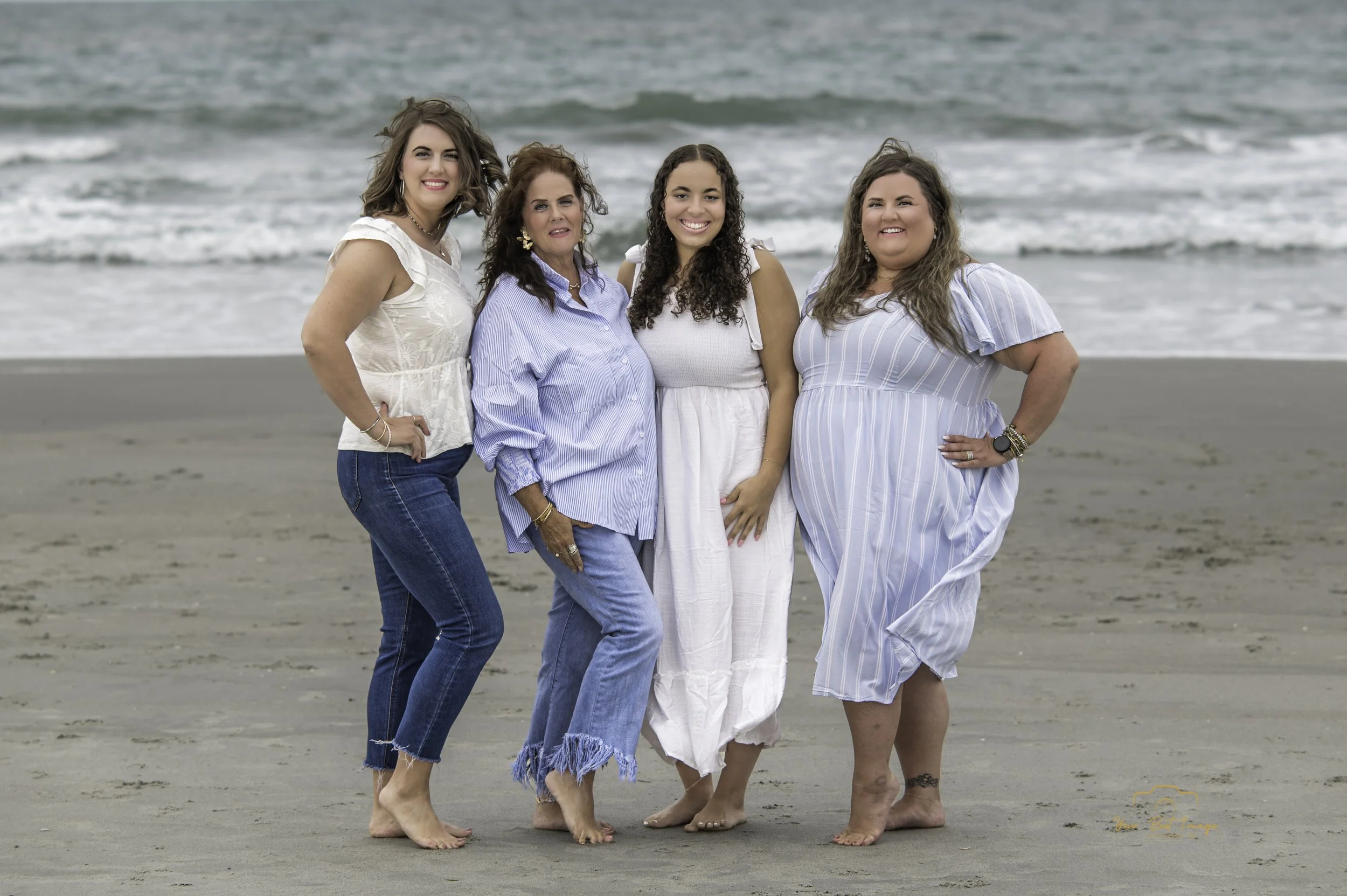 Four women standing barefoot on the beach near the ocean, smiling at the camera, dressed in light, casual clothing for warm weather.