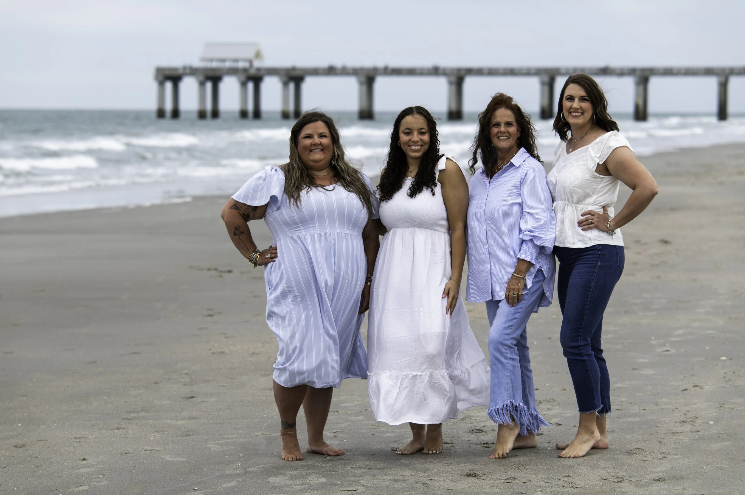 Four women standing on a beach near the ocean with a pier in the background. They are smiling and dressed in white and blue clothing, with some barefoot.