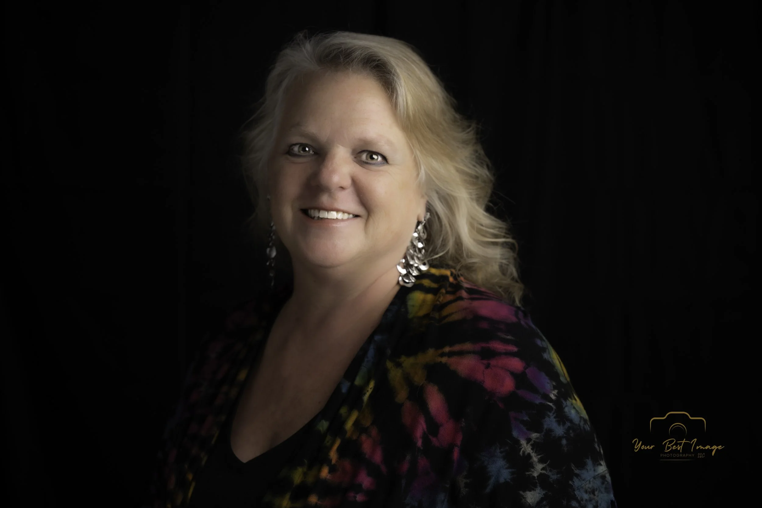 A woman with curly blonde hair smiling, wearing a colorful patterned top with shiny earrings, on a black background.