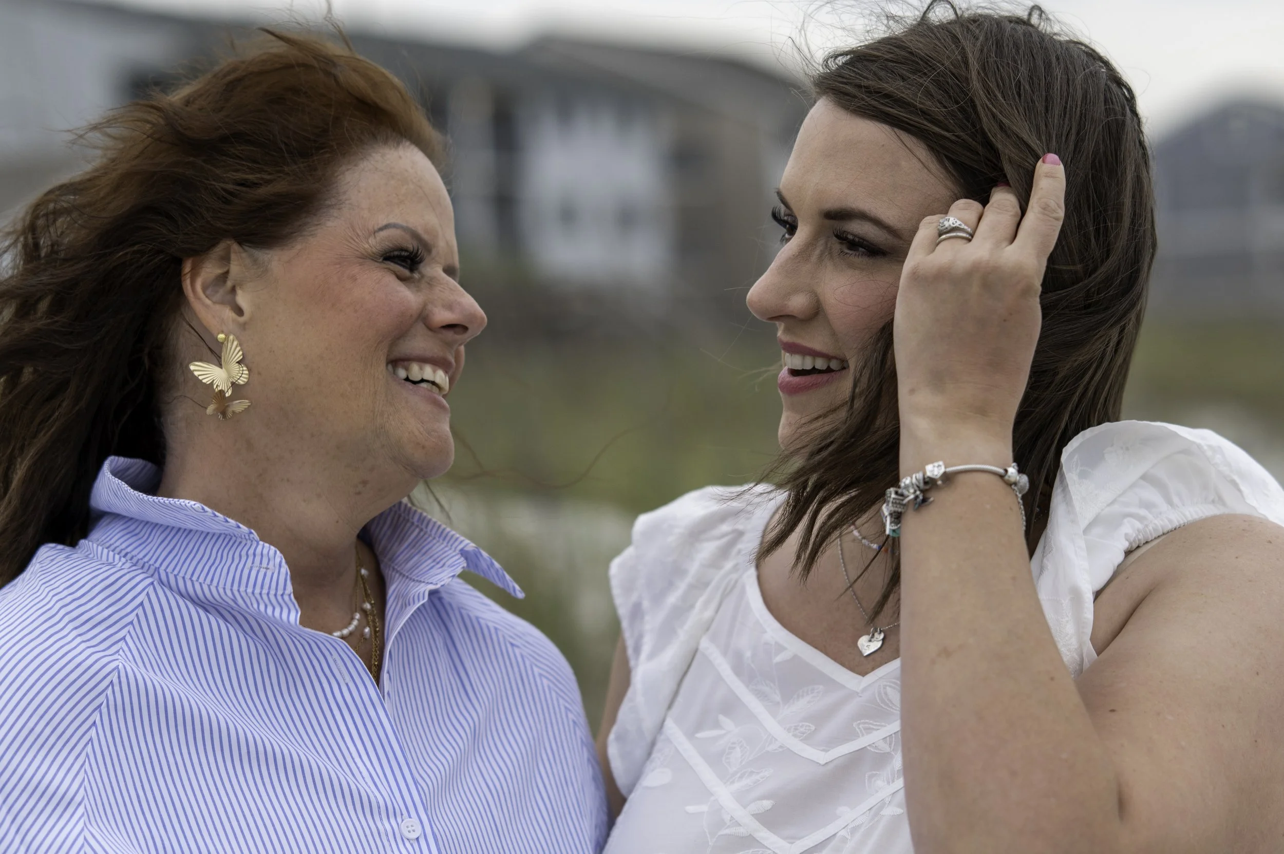 Two women smiling and looking at each other, one has red hair and butterfly earrings, the other has brown hair and is wearing jewelry and a white top.