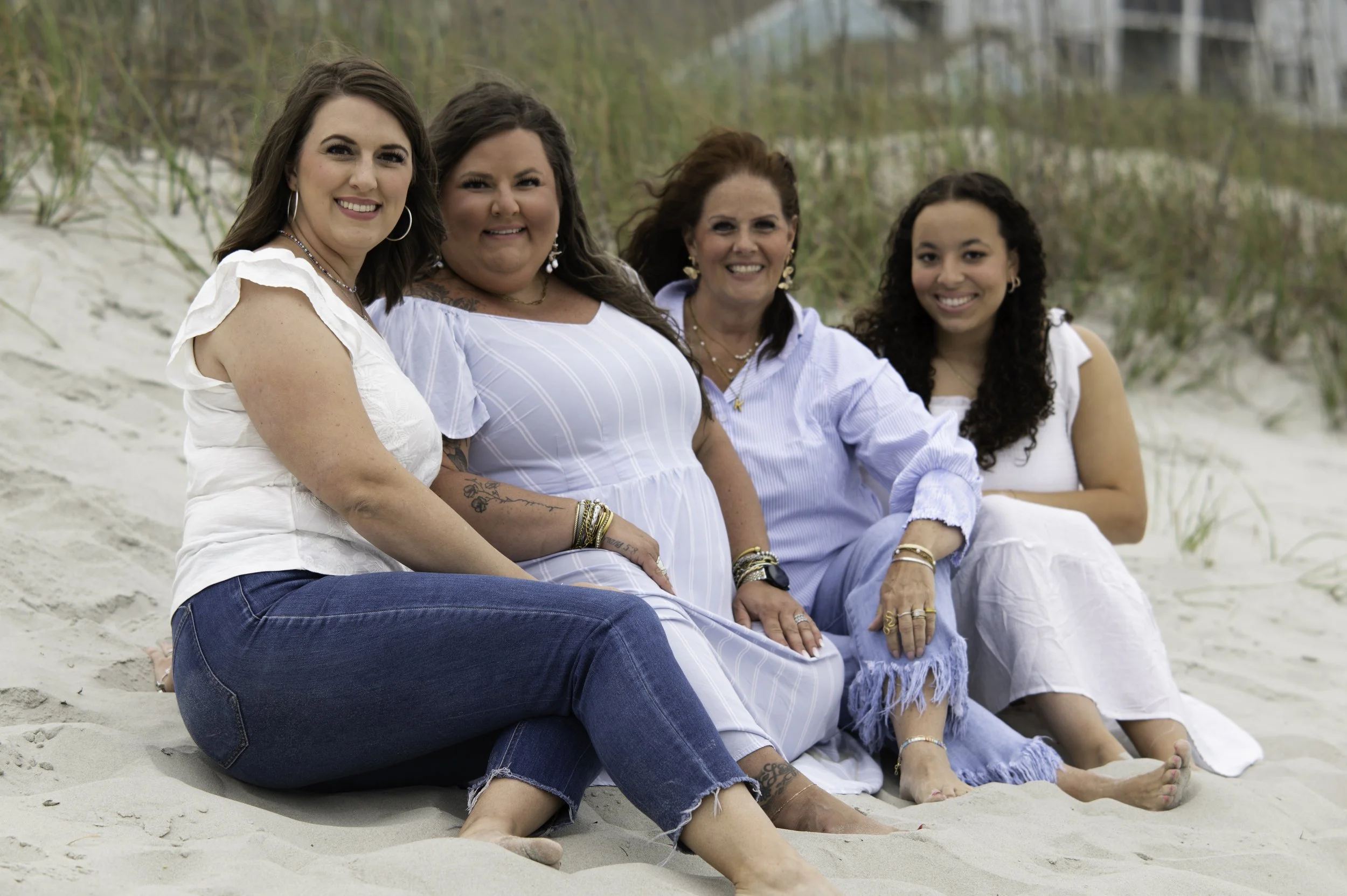 Four women sitting on the sands of a beach, smiling at the camera with a background of beach grass and buildings in the distance.
