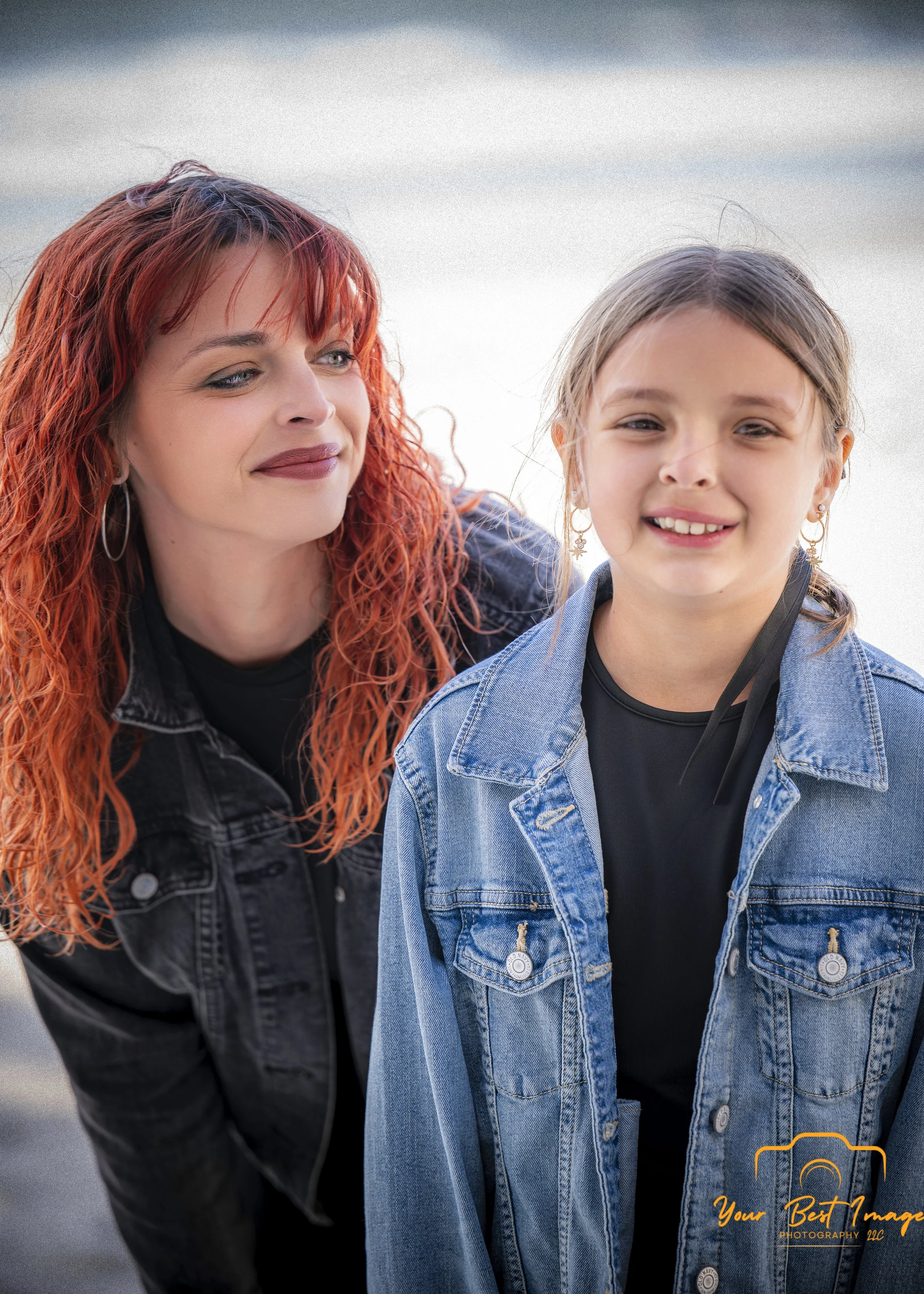 A woman with curly red hair and earrings looking at a girl with straight brown hair, earrings, and a denim jacket, both smiling. The background is blurred.