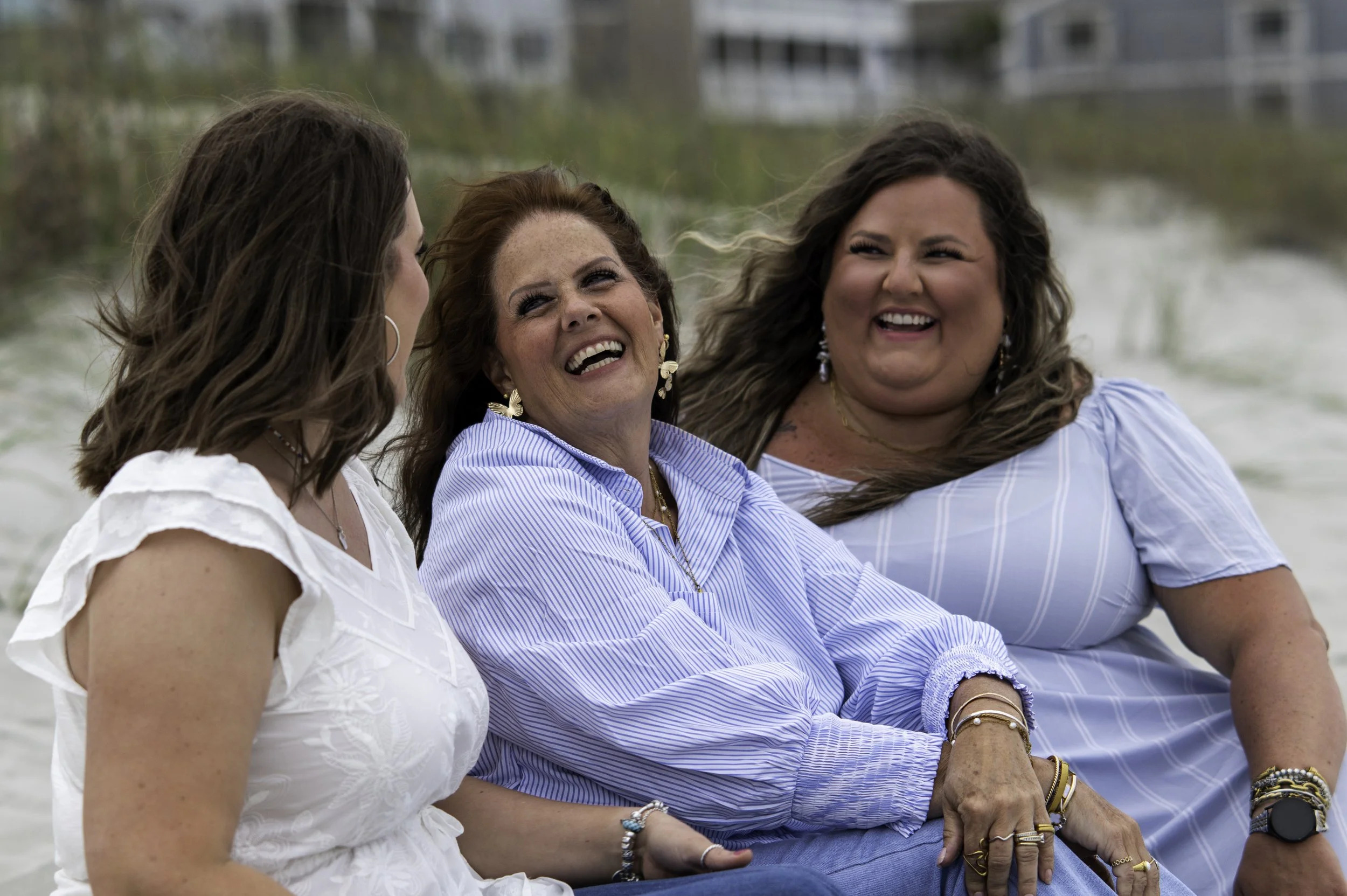 Three women sitting outdoors near water, smiling and laughing together.