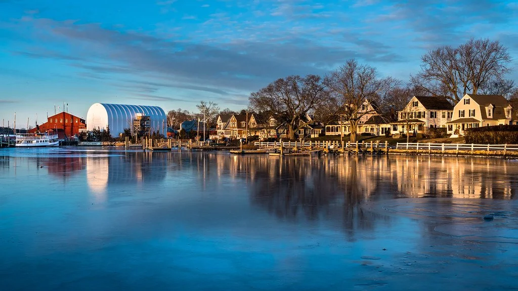 A serene harbor scene with calm water reflecting houses, trees, and a cloudy blue sky, featuring a large white storage building and boats.