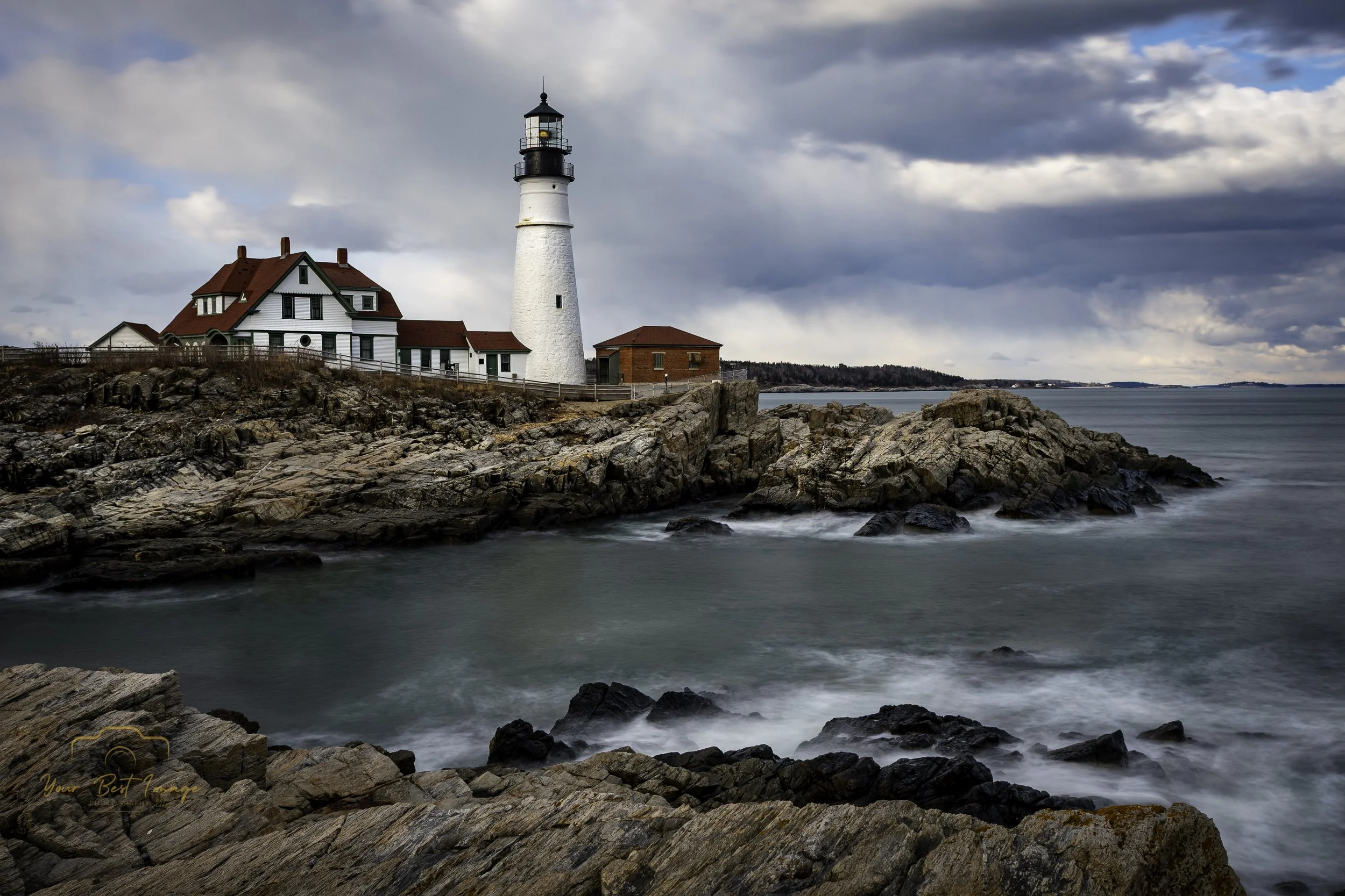 A lighthouse with a white tower and black top, accompanied by a white house with a red roof, sitting on rocky coast near the ocean under cloudy sky.