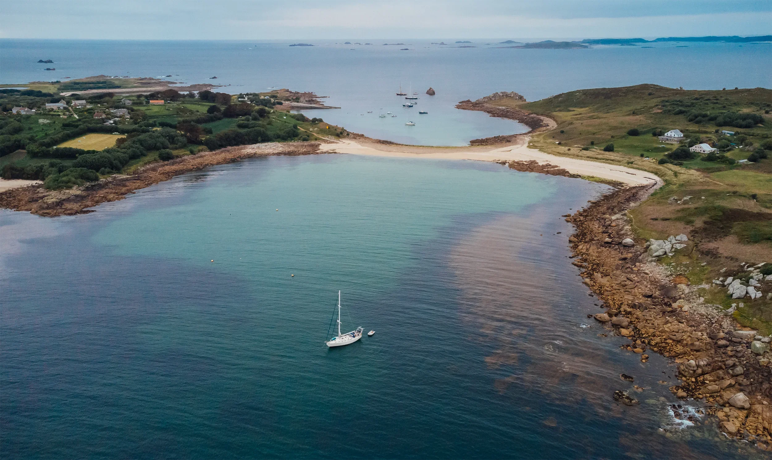 Aerial view of a coastal Scilly Isles bay with a yacht anchored near rocky shoreline, surrounding green hills