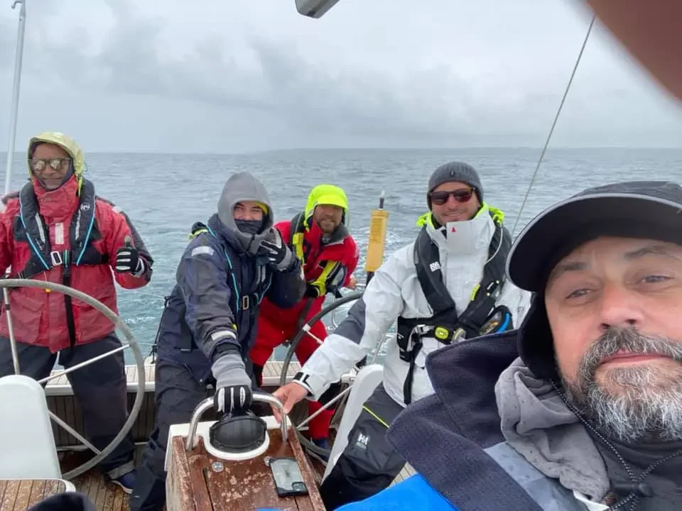 Sailing group in rain gear on a yacht with open ocean in the background, taking a group selfie.