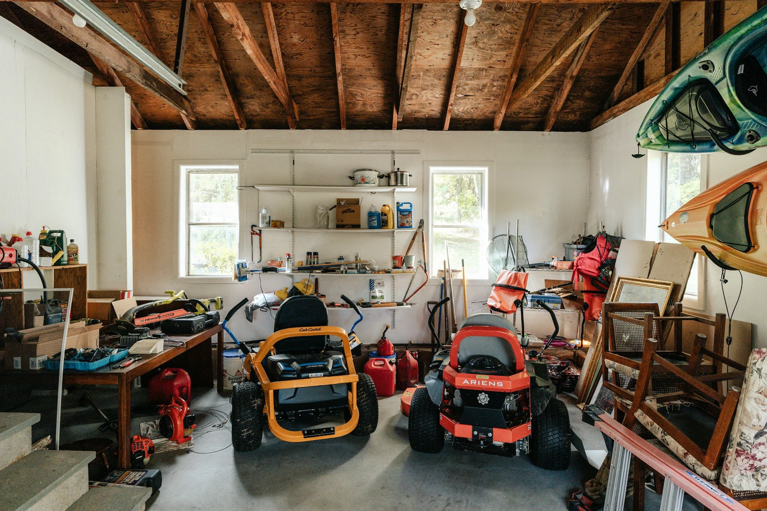 A cluttered garage with various tools, gardening equipment, and two riding lawn mowers. Kayaks are hanging on the right wall, and there are shelves and various items scattered on a workbench and around the room.