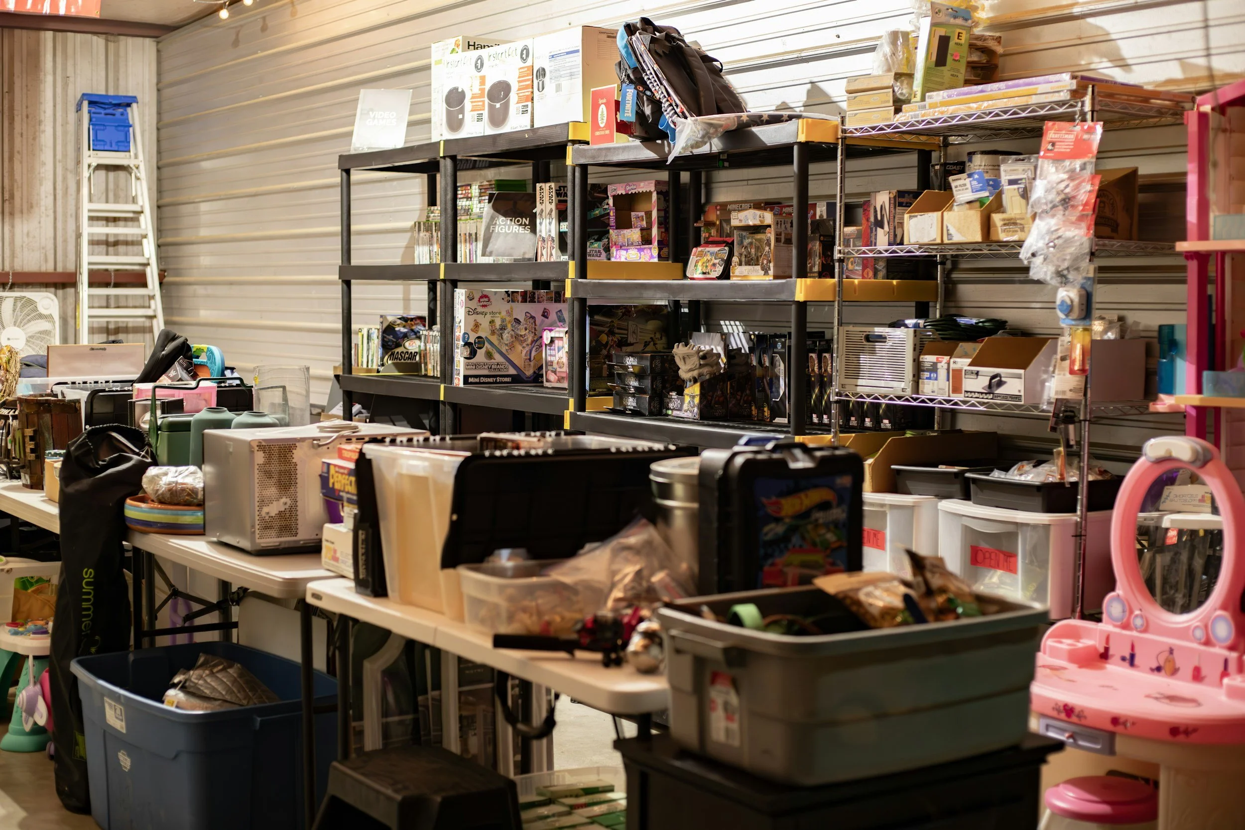 Shelving unit with toys and boxes, tables with various household items, and a plastic pink girl’s vanity with mirror in a storage unit or garage.