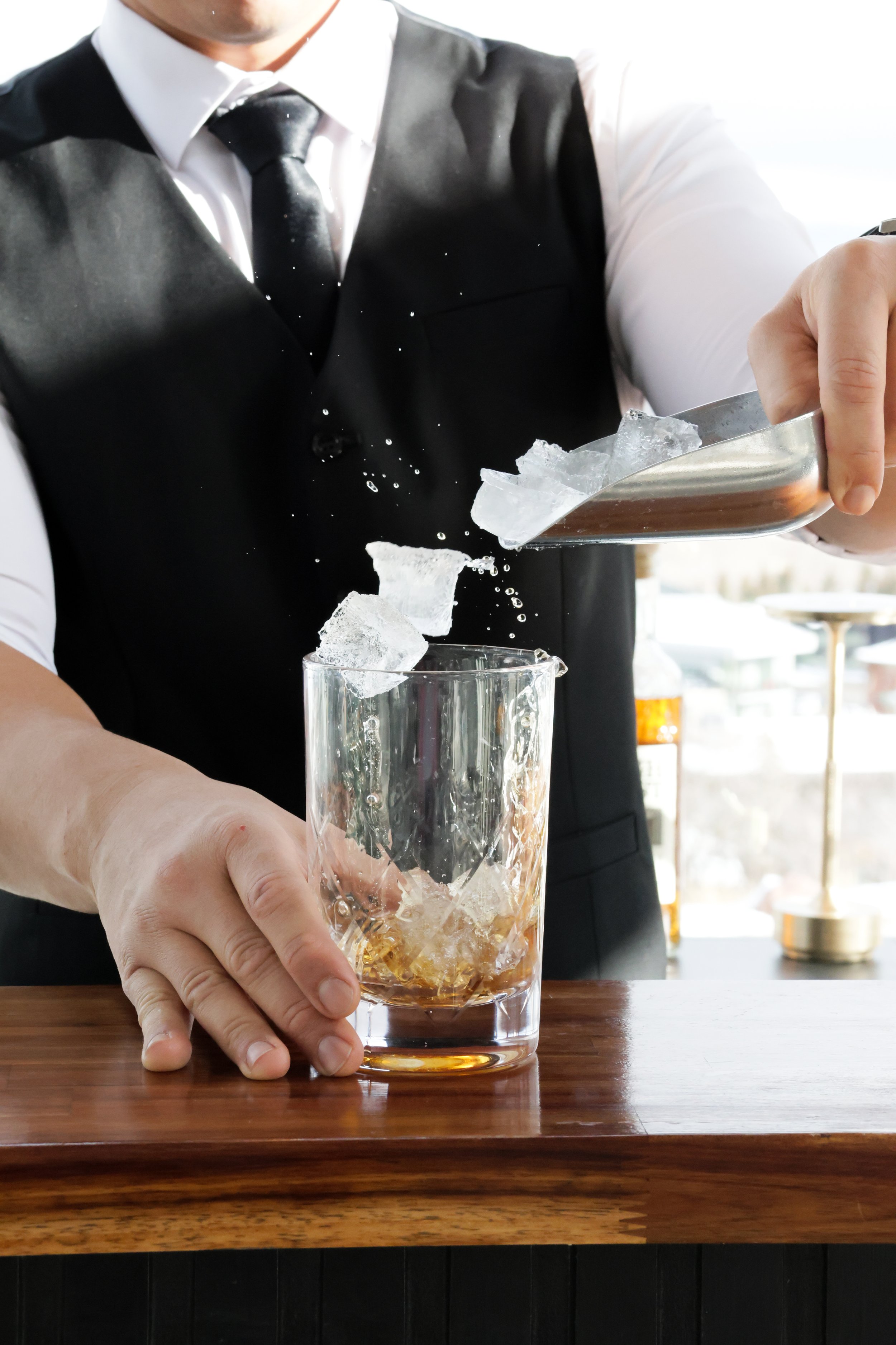 A bartender in a black vest, white shirt, and tie is pouring ice into a glass filled with whiskey on a wooden bar counter.