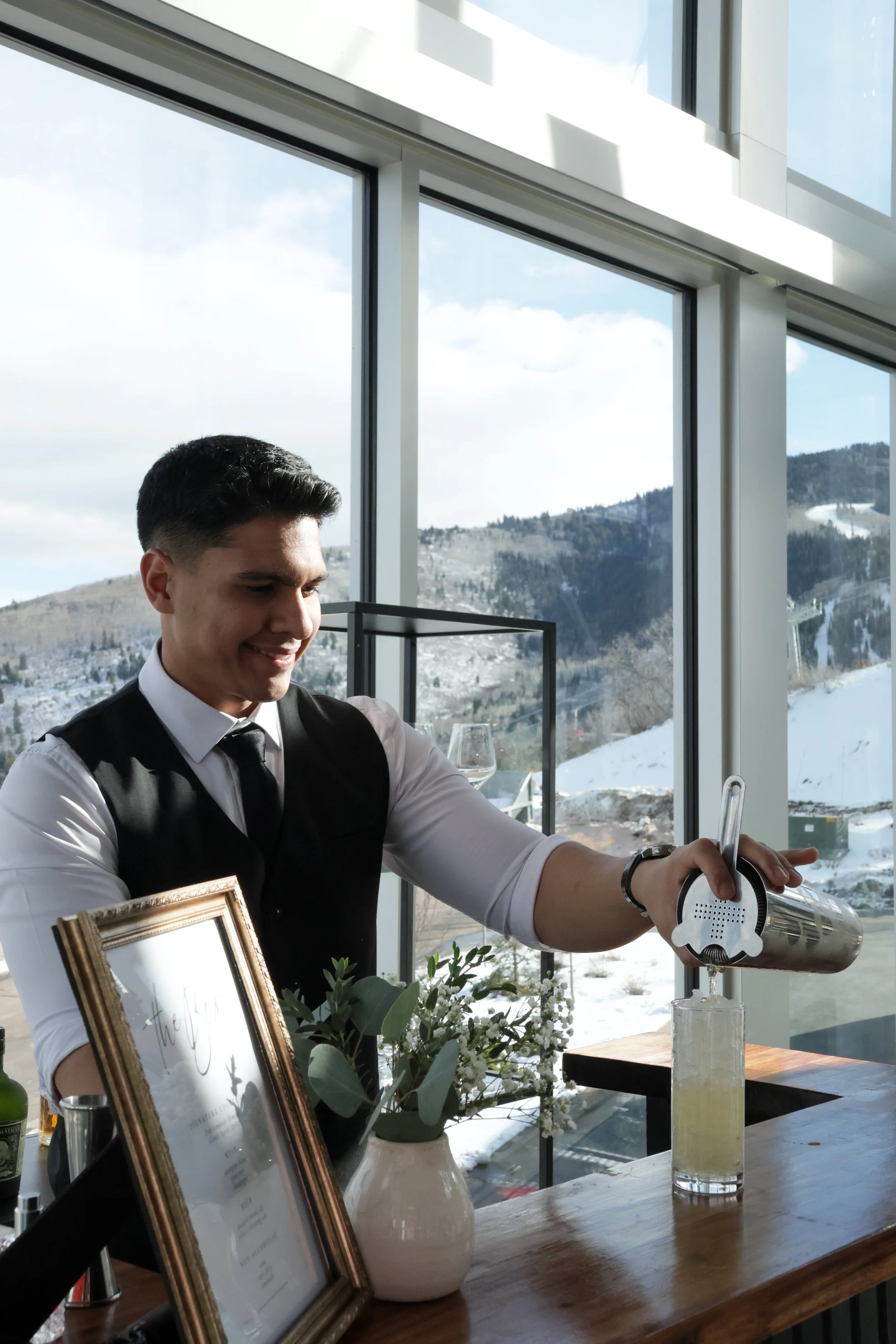 A bartender in a white shirt and black vest making a drink with a handheld blender at a bar counter inside a restaurant with large windows and snow-covered mountains in the background.