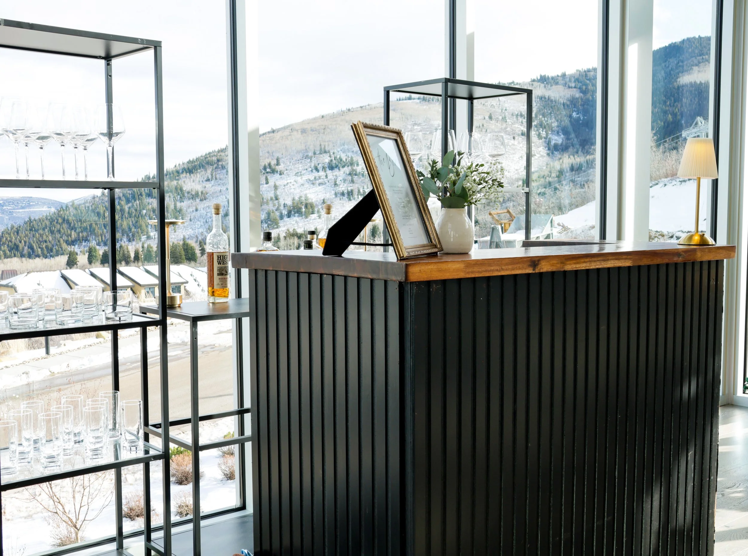 Interior of a modern bar with large windows showing snow-covered mountains outside, featuring a wooden bar counter, glass shelves with glasses, a framed menu, a bottle of whiskey, a potted plant, and a table lamp.