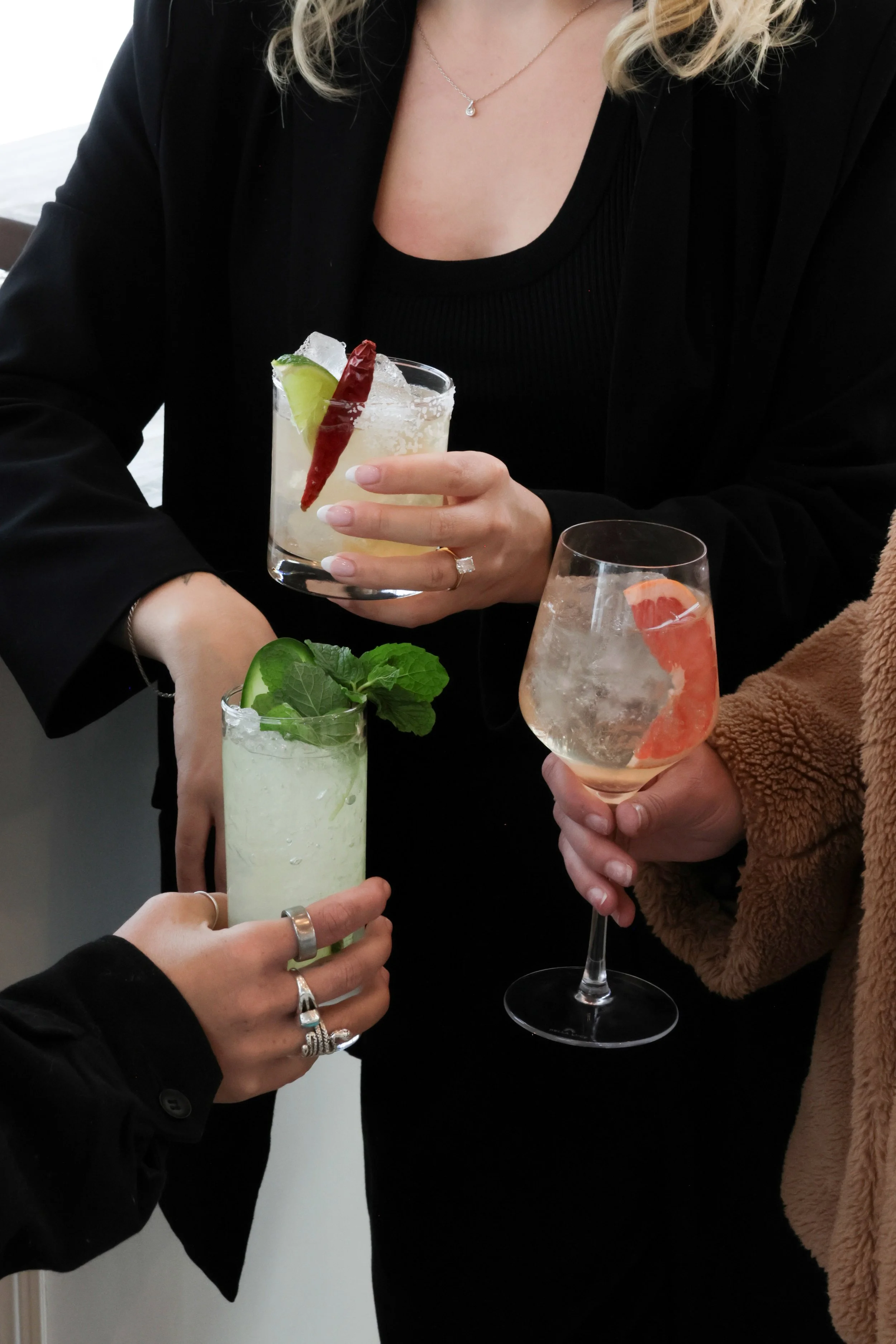Three women holding different cocktails for a toast, with only their hands and parts of their bodies visible.