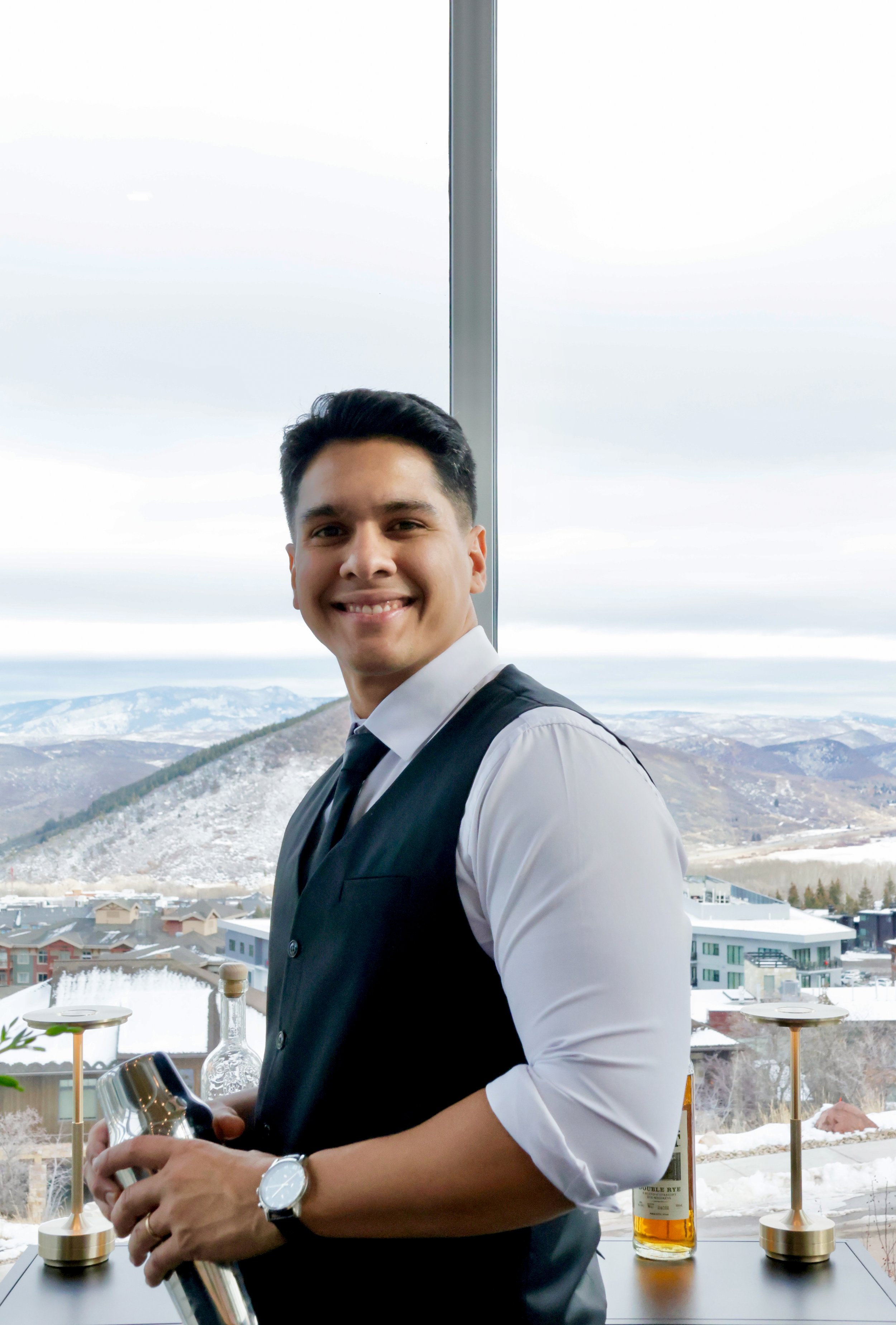 A smiling man in a tuxedo vest and white shirt standing in front of a window with a mountain and snowy landscape view, holding a cocktail shaker.