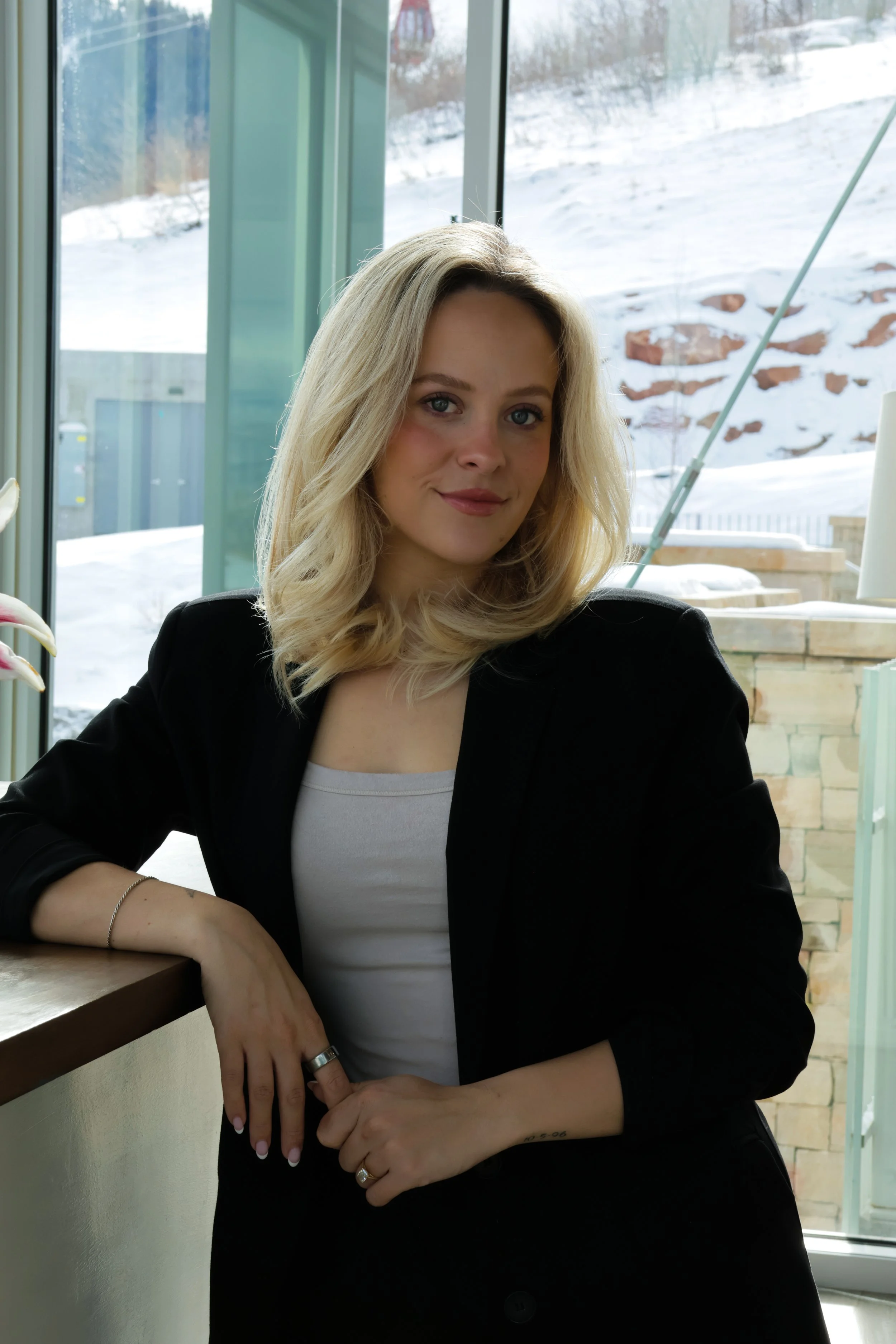 A woman with blonde, shoulder-length hair, wearing a black blazer and a white top, sitting indoors near a window with a snowy outdoor background.