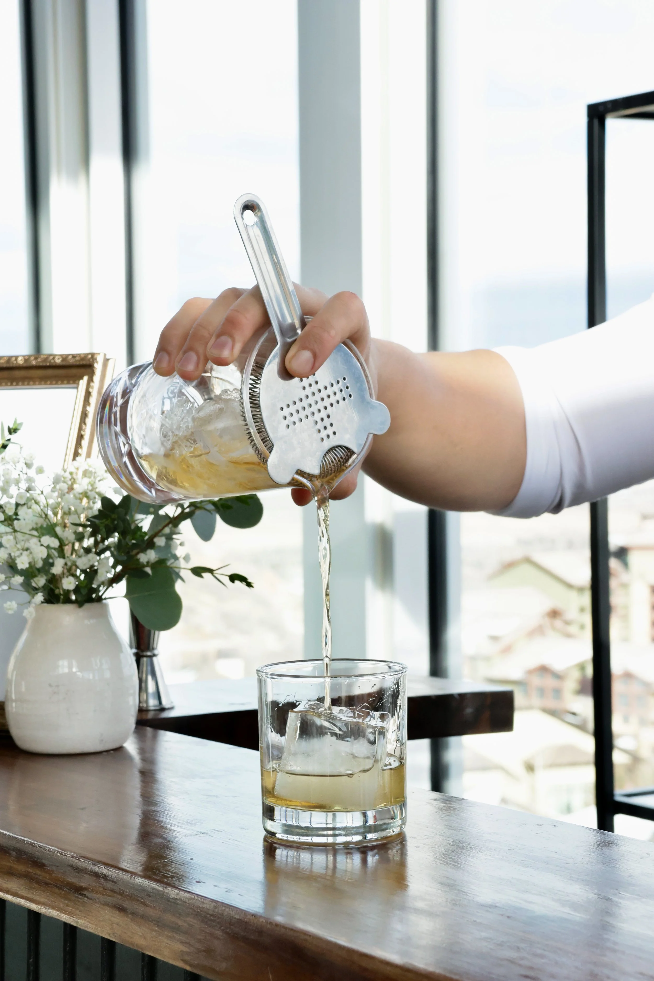Person pouring a drink with ice into a glass using a metal citrus juicer over a wooden table near a window and decorative flowers.