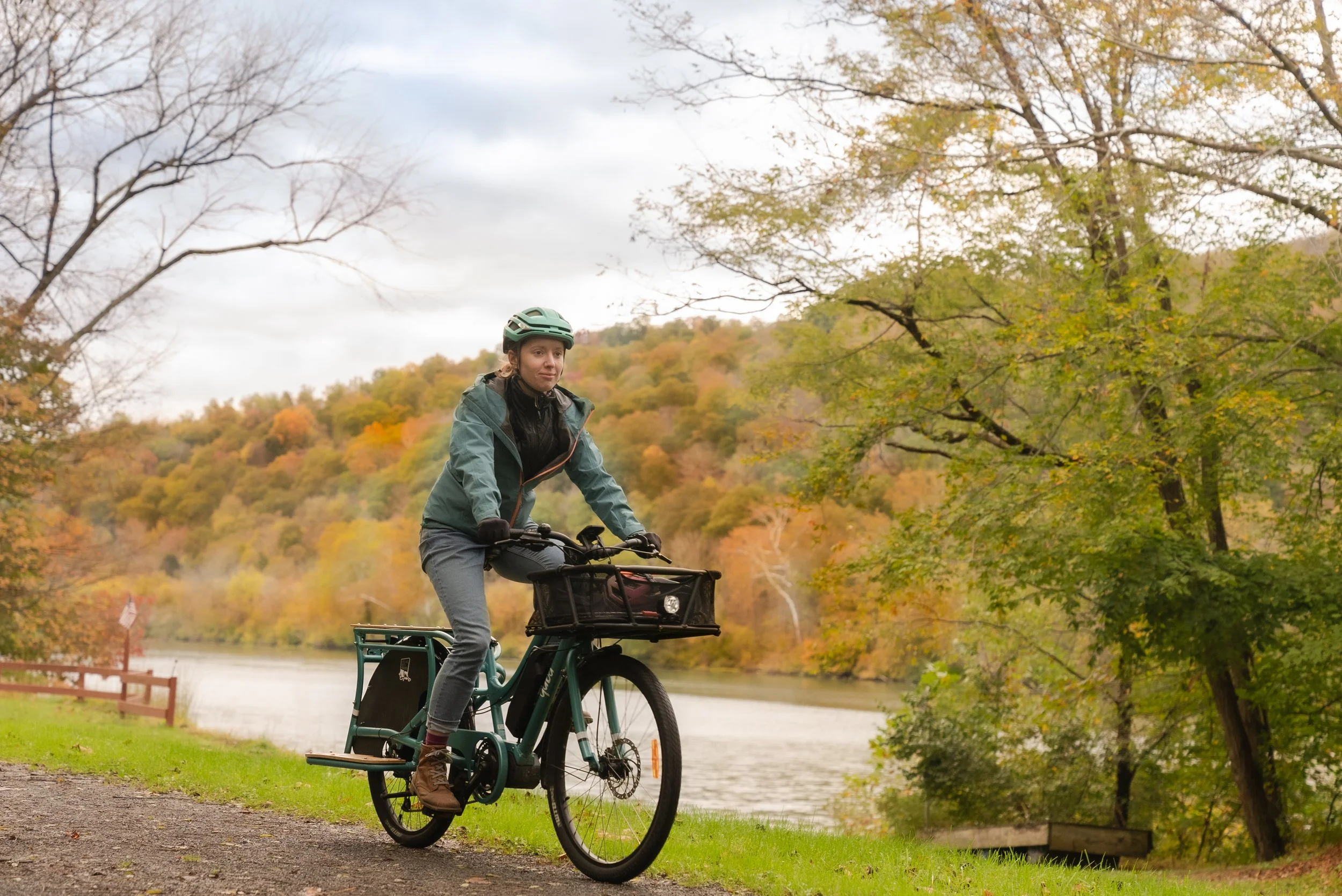 A woman riding an e-bike on a path beside a river during autumn, with trees displaying fall foliage and overcast sky in the background.
