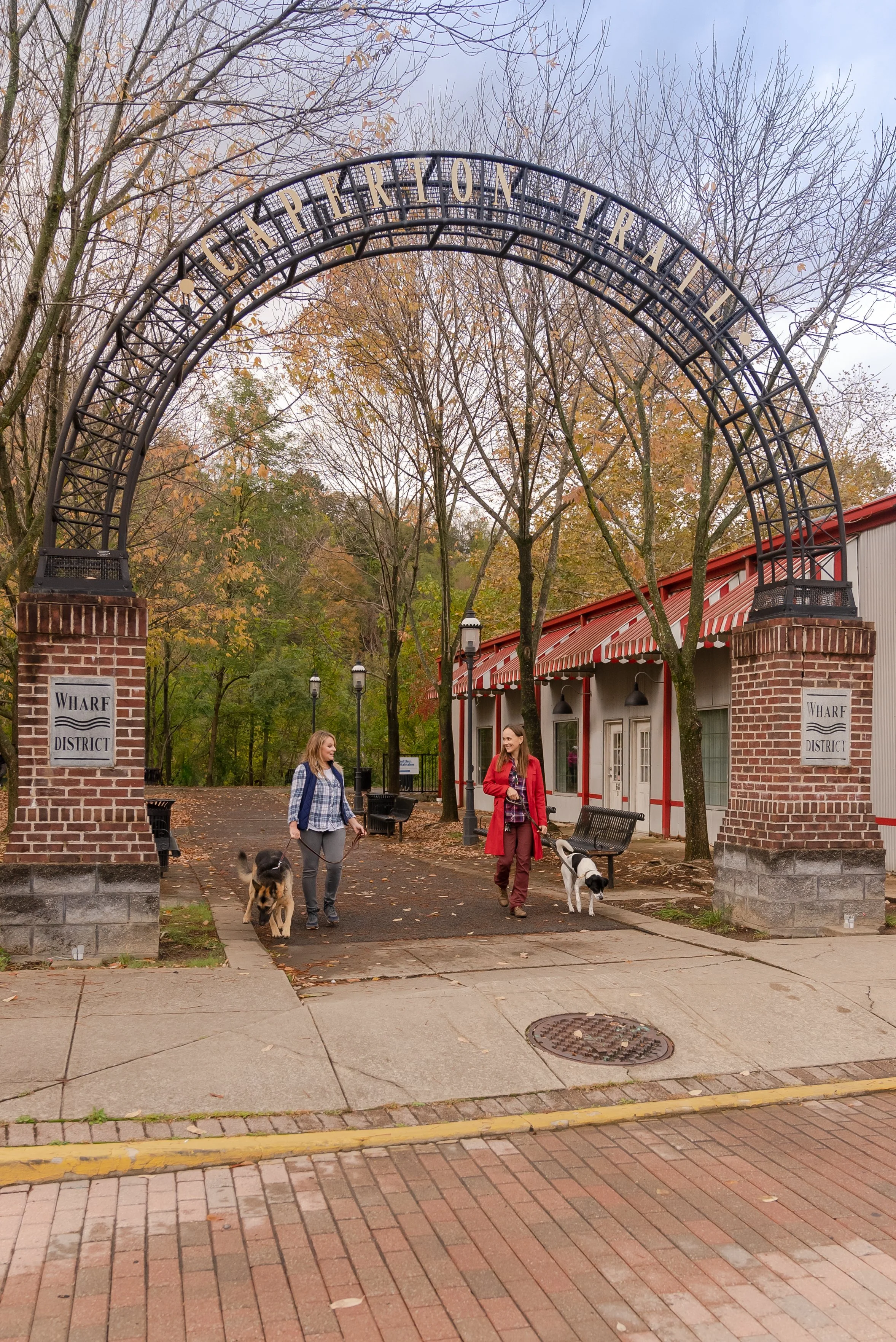 Two women walk dogs under an arched entrance gate that says 'Experience Trail' in a park setting with trees and pathway, near a red and white building.