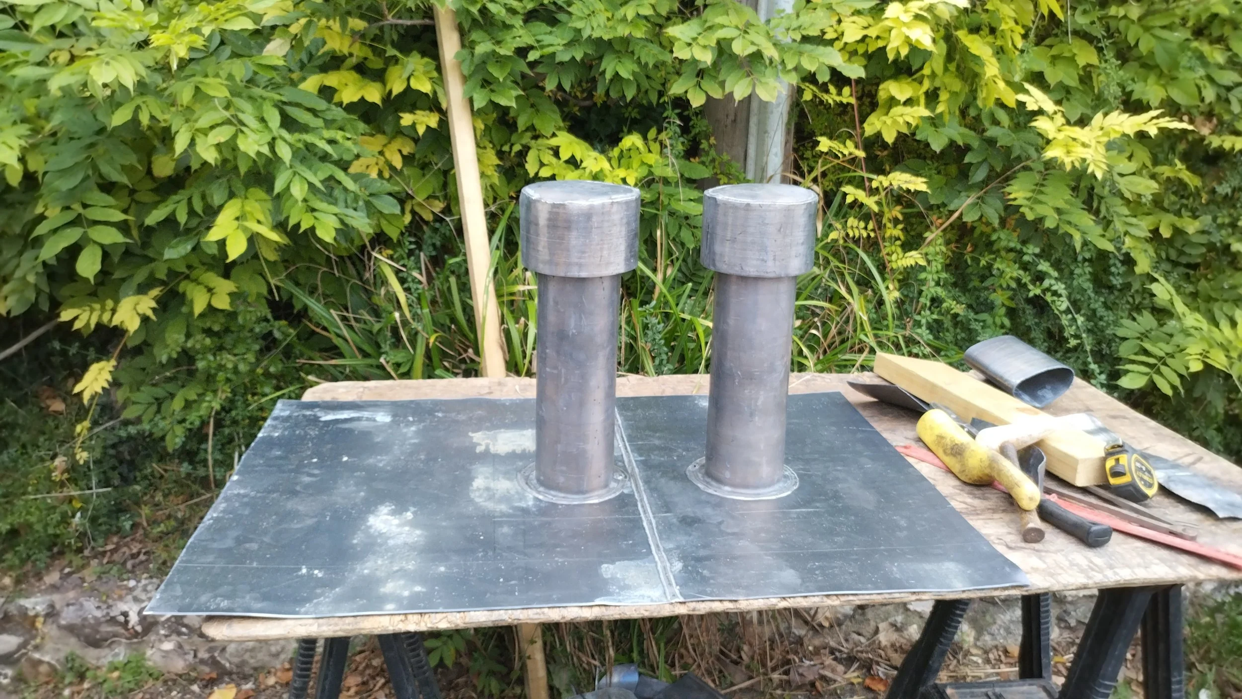 Metal pipes and tools on a wooden workbench outdoors, with green leafy bushes in the background.