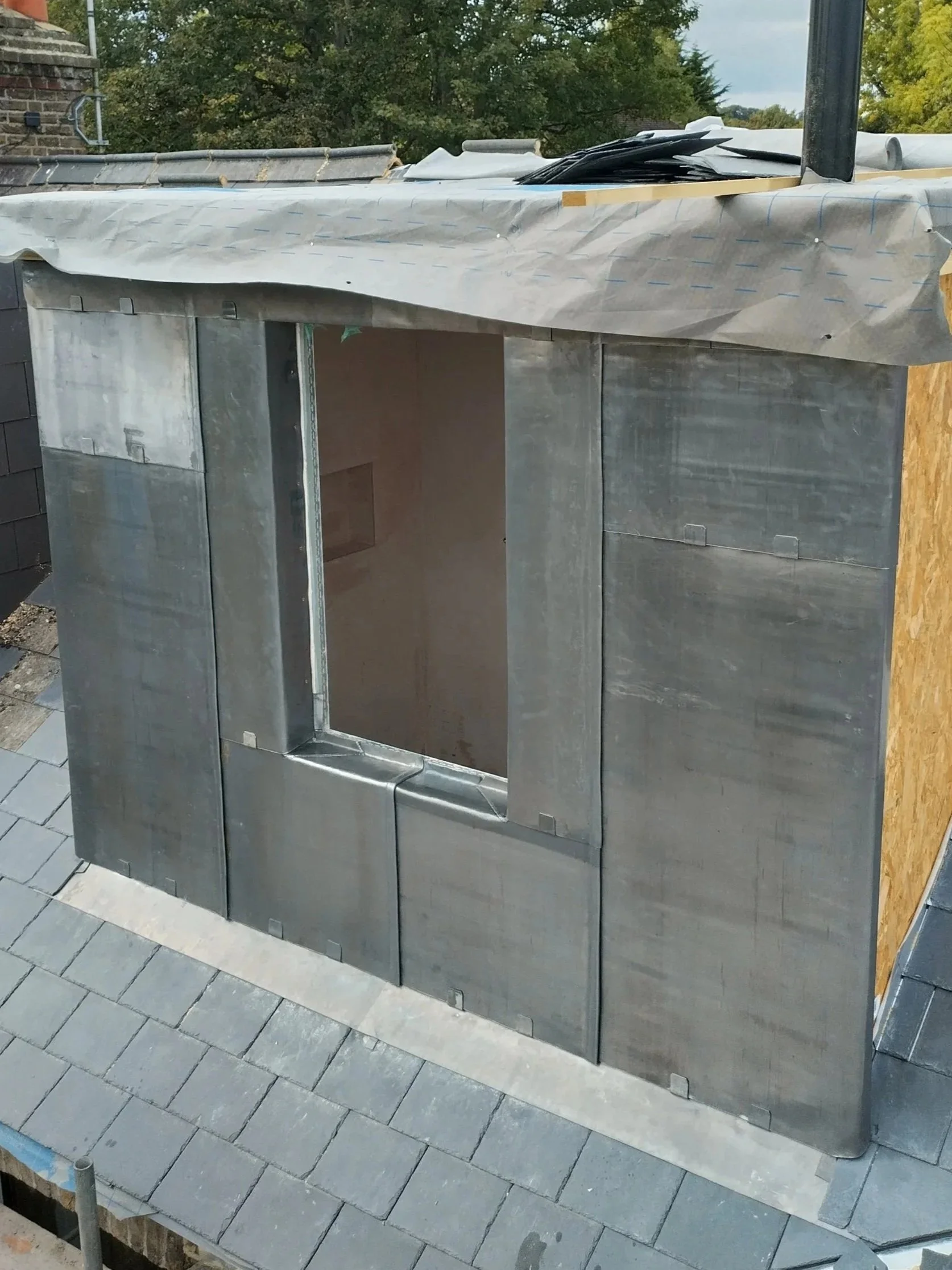Metal framework and insulation installed on a roof with shingles visible at the bottom, with a view of trees and a cloudy sky in the background.