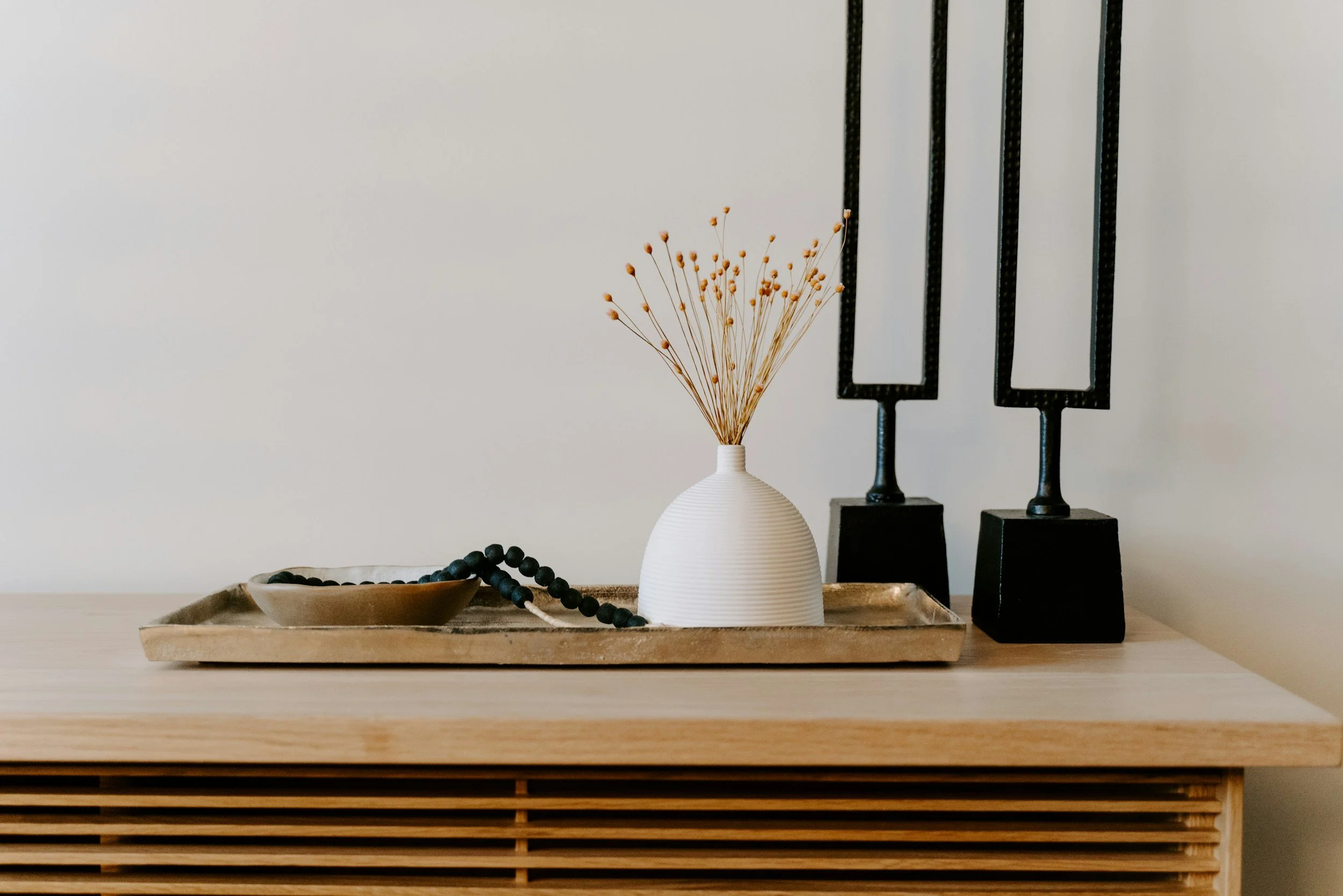 Decorative arrangement on a wooden surface with a small beige tray, a black beaded necklace, a white vase with dried flowers, and black sculpture pieces.