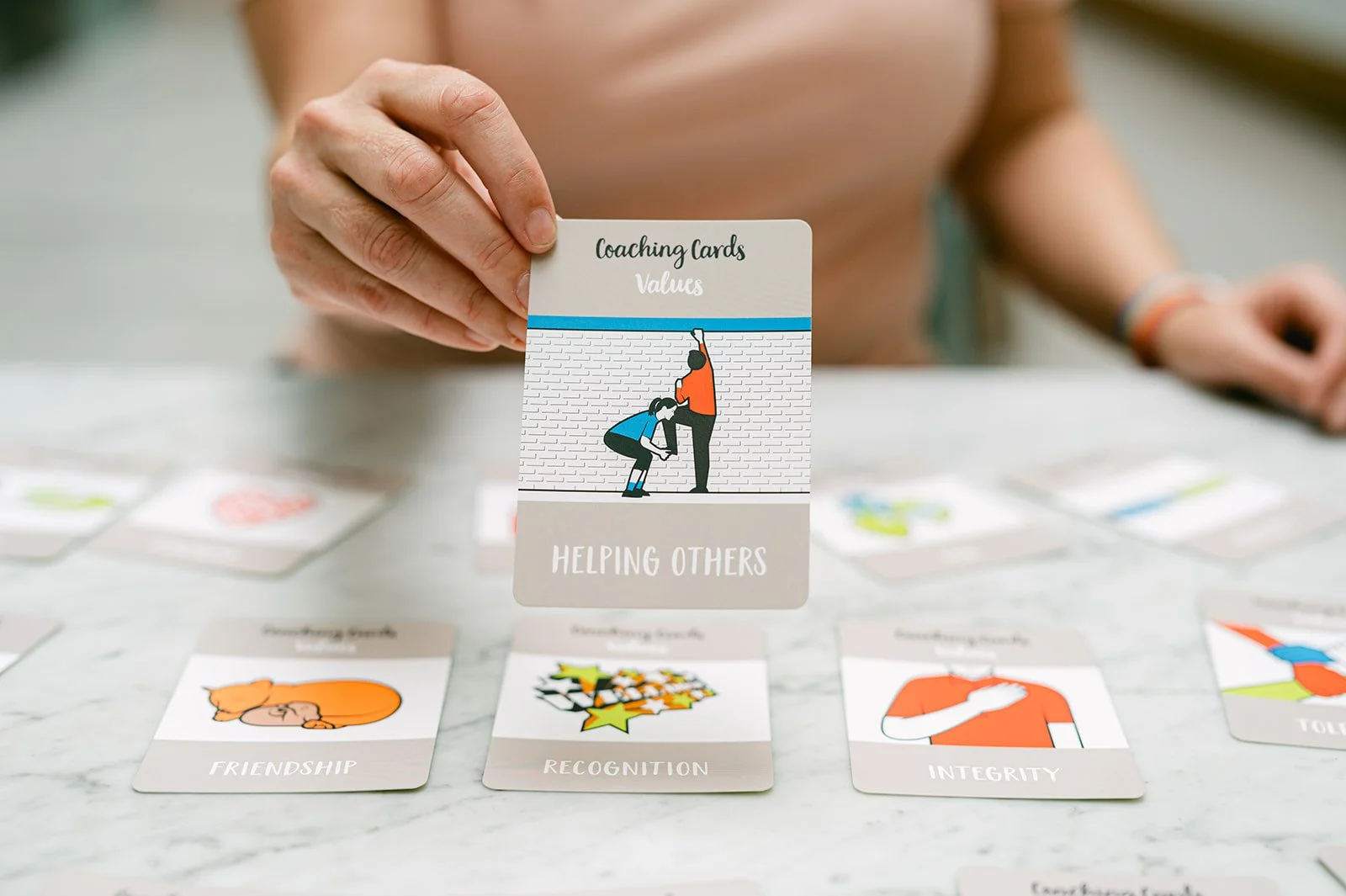 A table is covered in picture cards with one being held up. That card has "Values" at the top followed by a picture of someone giving someone else a leg-up and "helping others" written underneath.