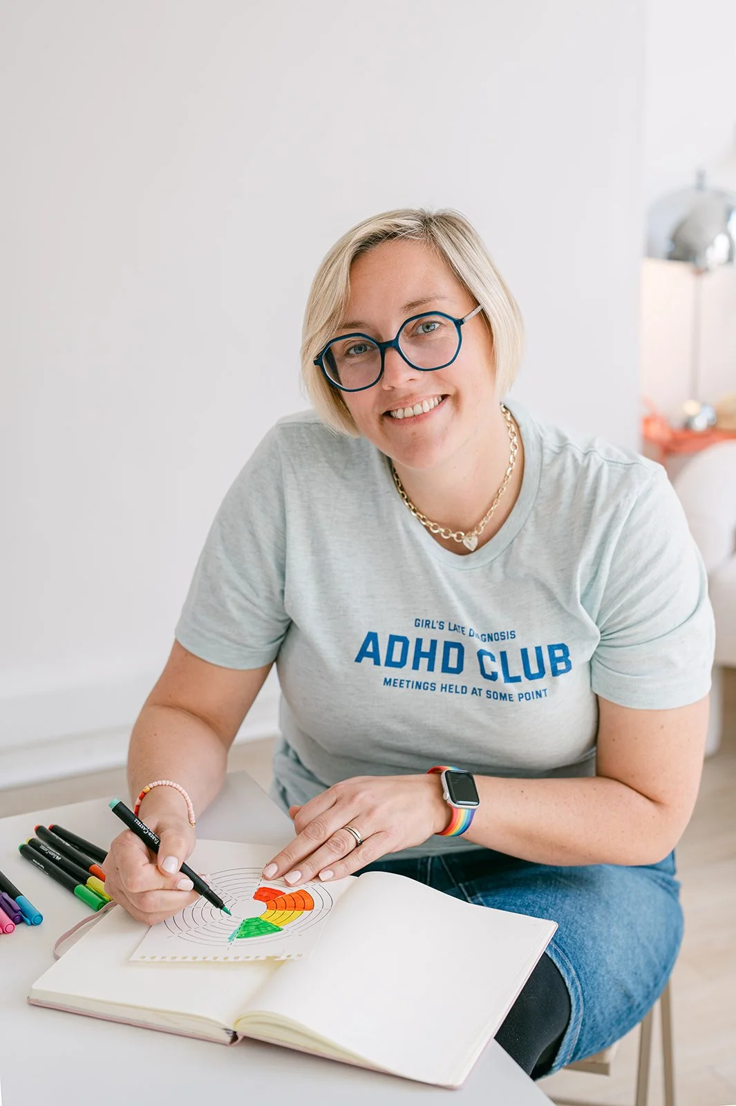 Portrait of Kate Faxen wearing a t-shirt that says "girl's late diagnosis ADHD club, meetings held at some point". She has a short blond bob and wears hexagonal turquoise glasses. She's colouring a wheel of life coaching exercise.