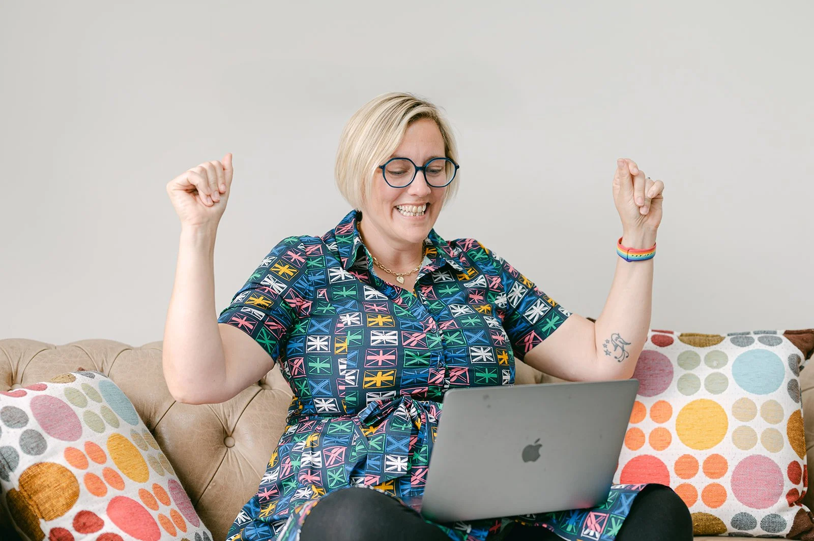A picture of Kate with her arms raised as if cheering. She's sat cross legged with her laptop on her knee and is wearing a dress covered in pink, yellow, blue and green union flags.