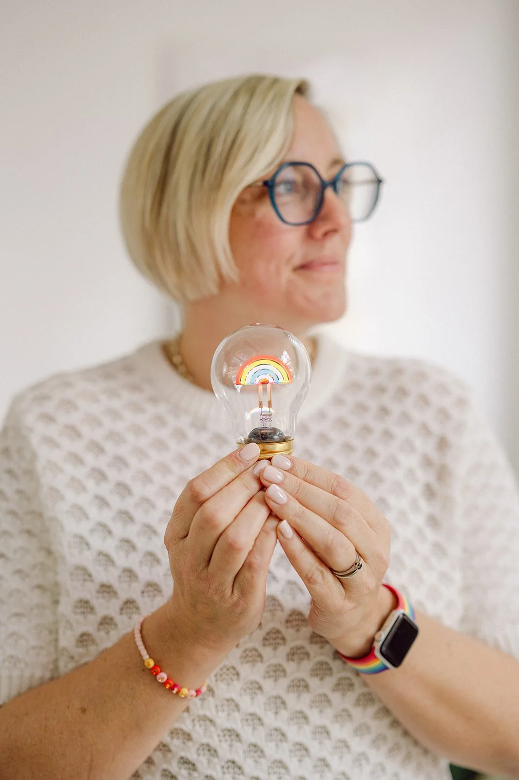 Kate is out of focus behind her hands which are in focus and holding a lightbulb with a rainbow inside.