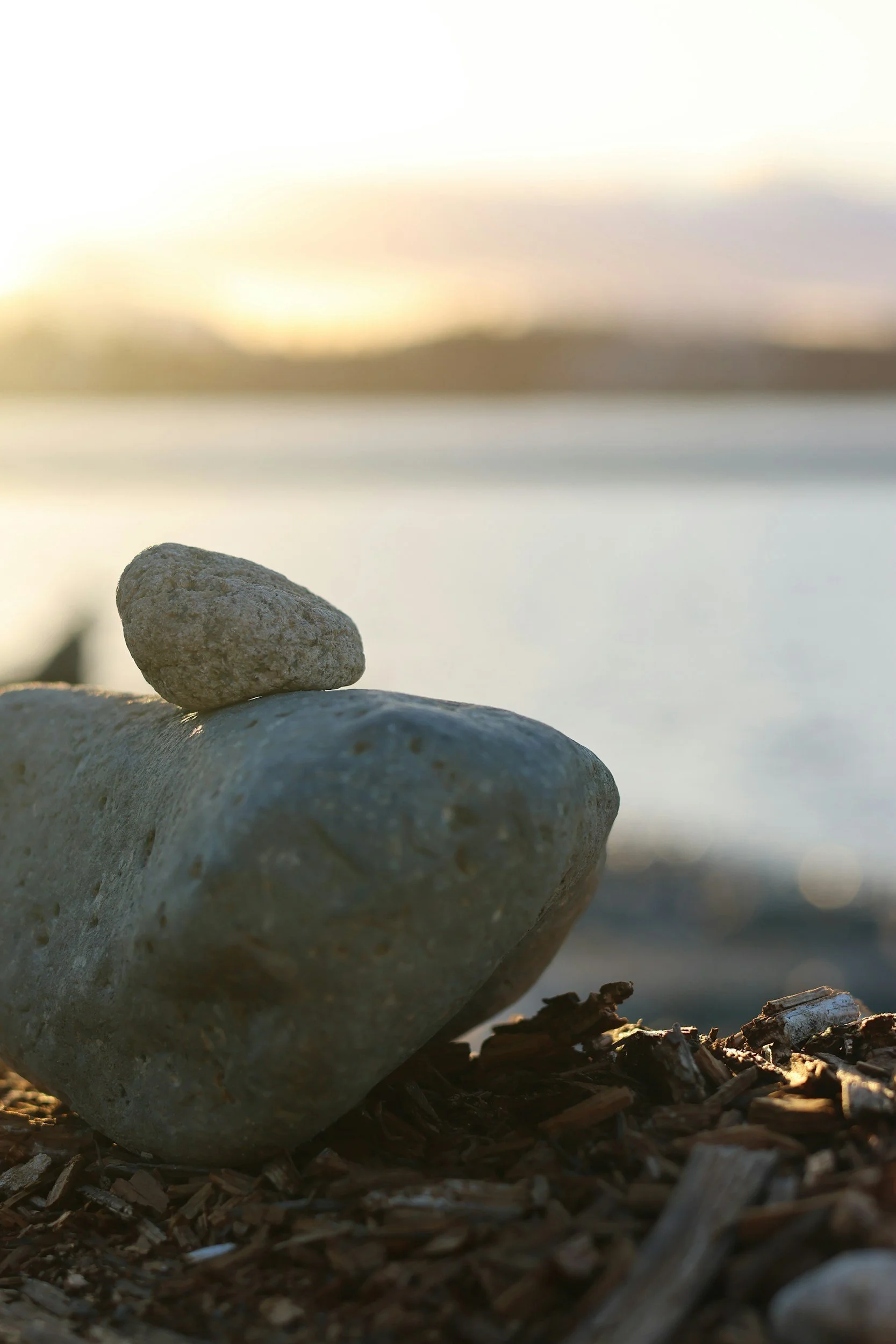 Close-up of two small rocks balanced on larger rocks on a shoreline with water and a blurred sunset in the background.