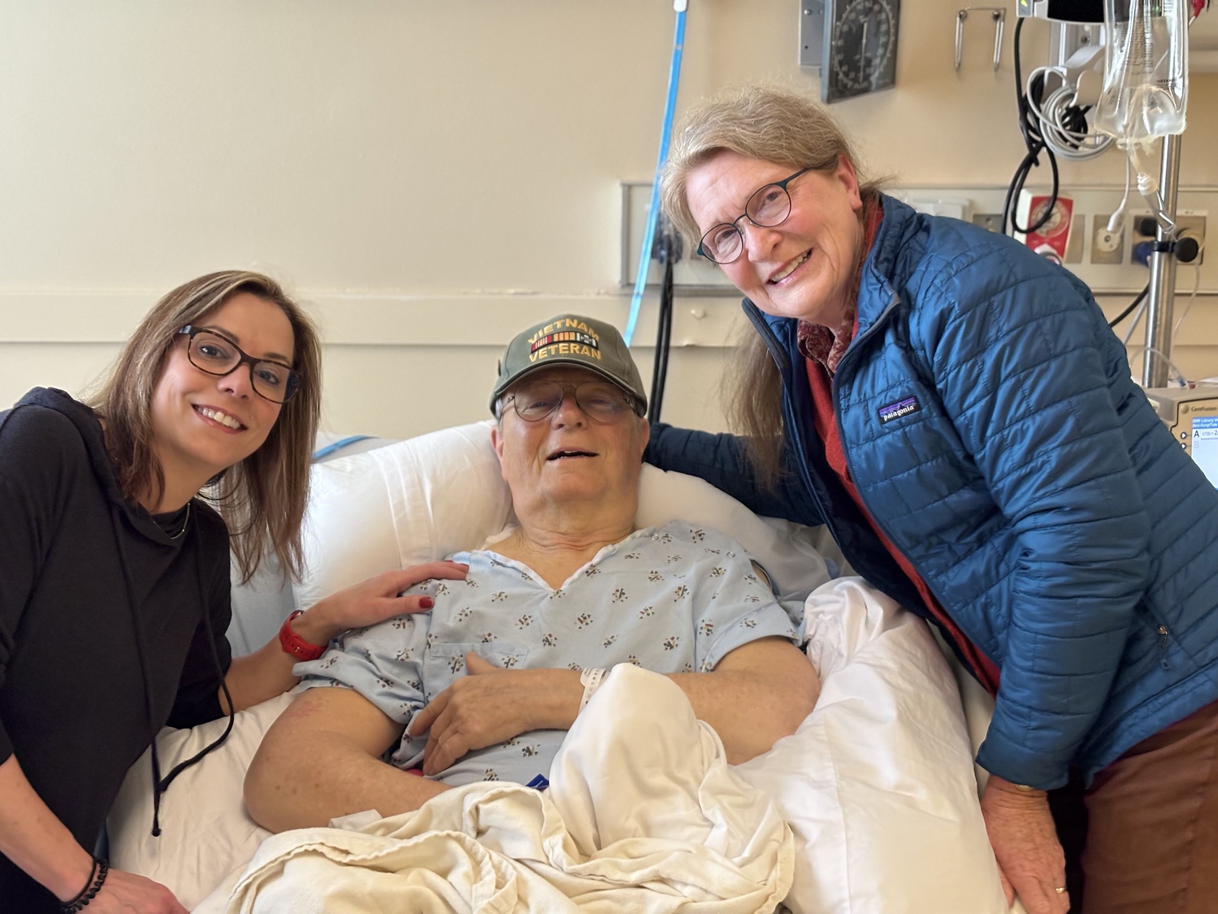 A man in a hospital bed wearing a cap with Vietnam veteran patches, smiling and surrounded by two women, one on each side of the bed, in a hospital room.