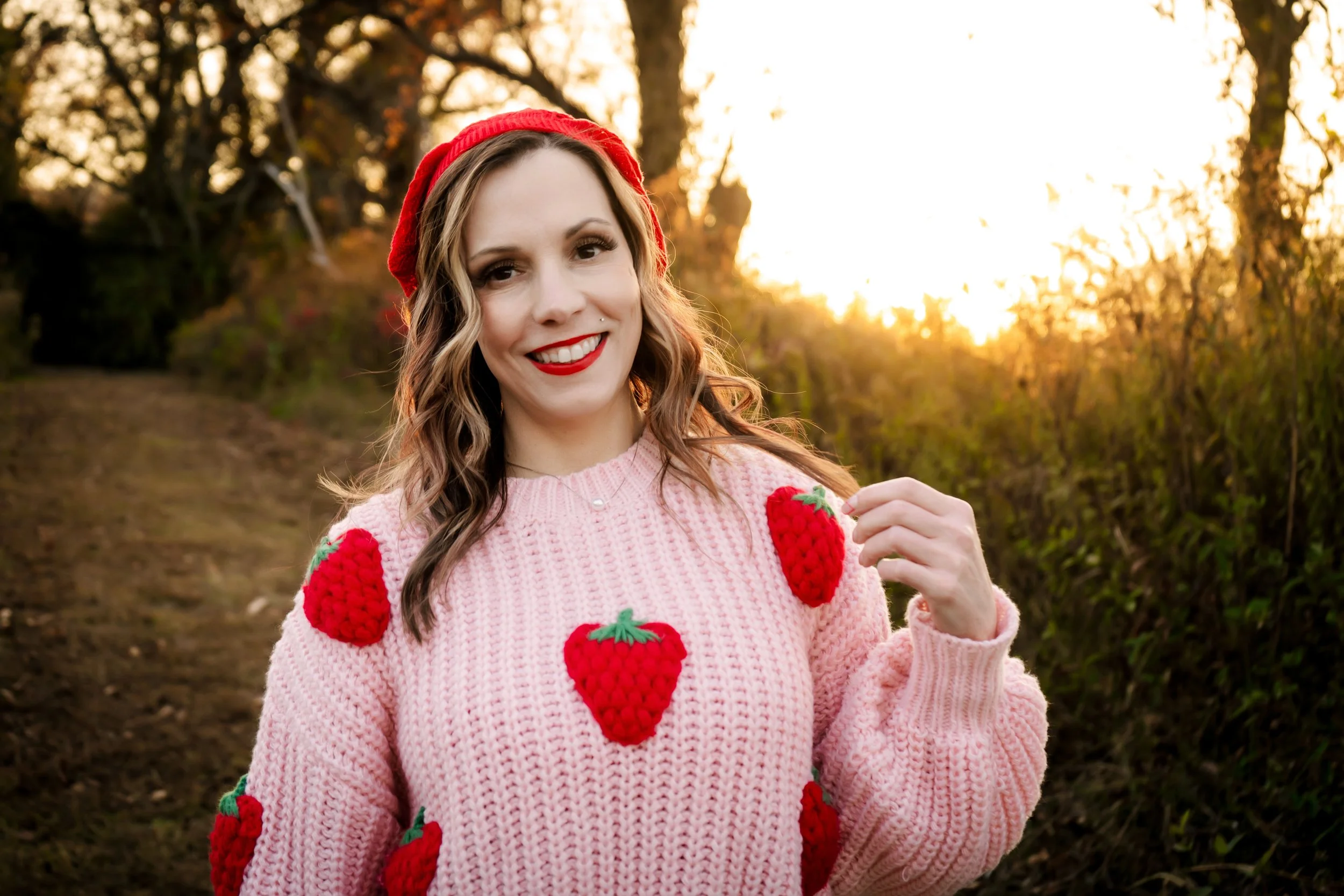 A woman outdoors at sunset, wearing a pink sweater with red strawberry patterns, a red beret, and red lipstick, smiling and touching her sweater.