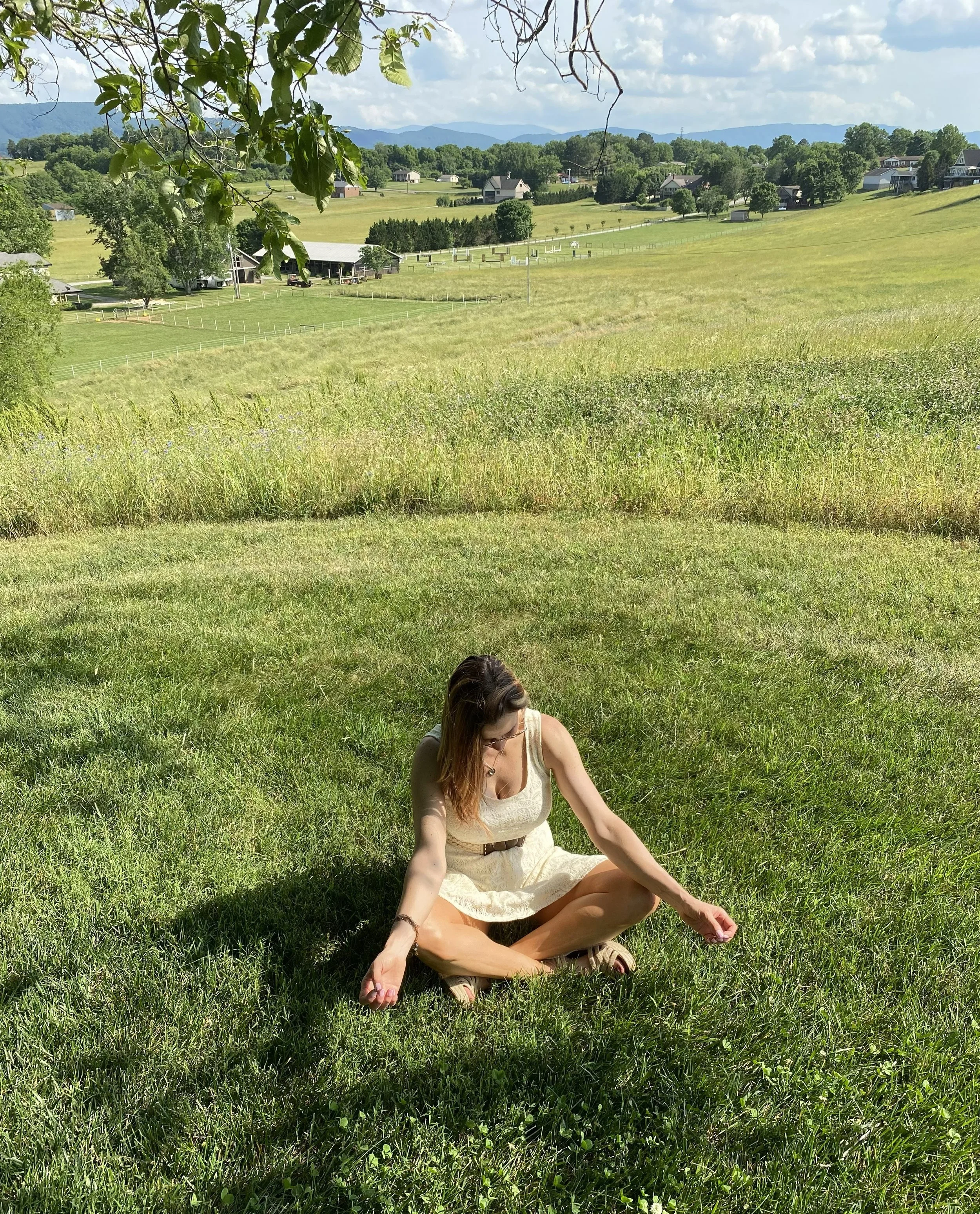 Woman practicing yoga or meditating on green grass in a sunny field with rolling hills and houses in the background.
