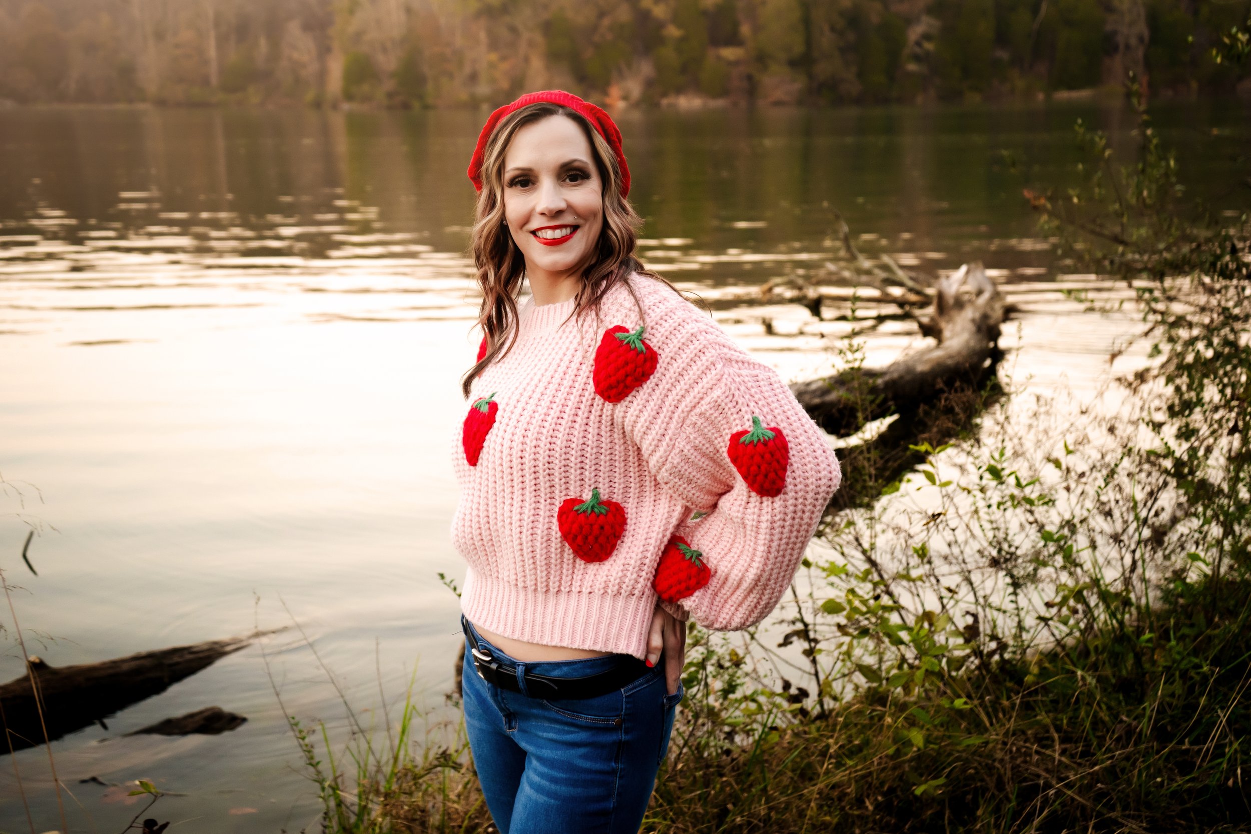 A woman wearing a pink sweater with red strawberry embellishments, a red headband, and blue jeans stands near a lake during sunset with trees in the background.