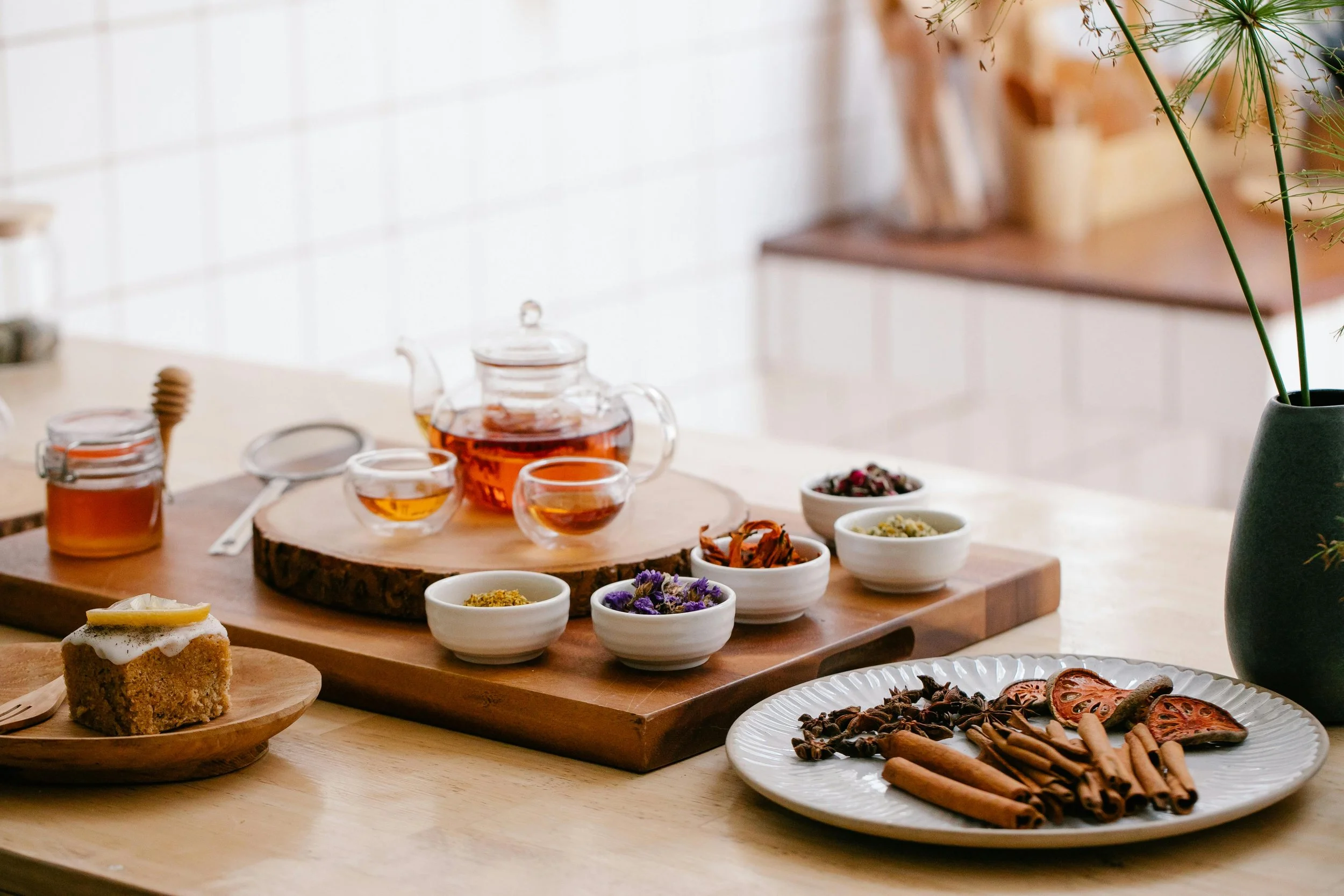 Tea party setup with a glass teapot, tea cups, bowls of herbs and spices, a slice of cake with lemon, and various dried herbs and spices on a white plate, all arranged on a wooden surface.