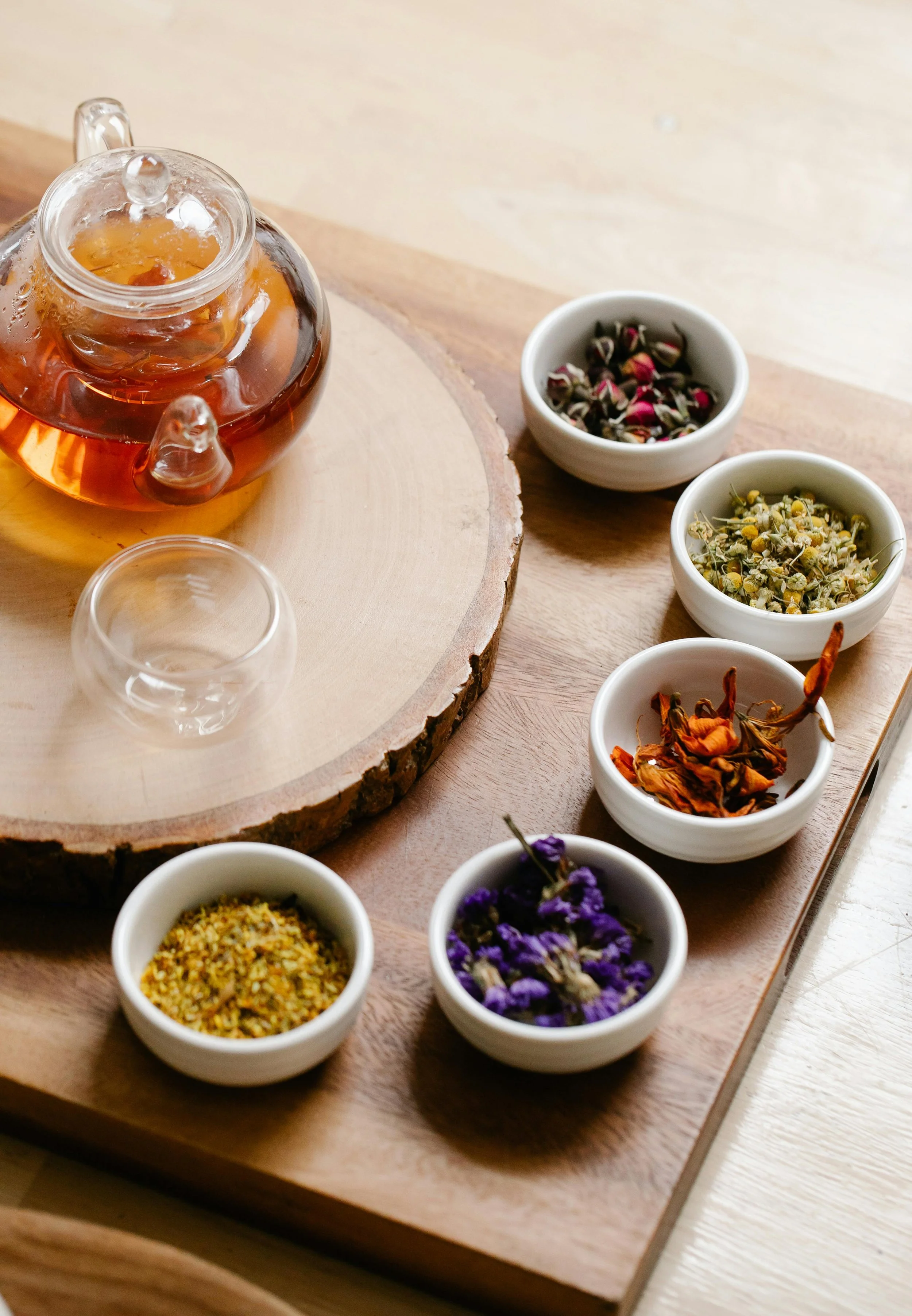A glass teapot filled with amber-colored tea resting on a round wooden serving board. Surrounding the teapot are small white bowls containing various dried flowers and herbs, including chamomile, hibiscus, lavender, and other colorful plant materials.