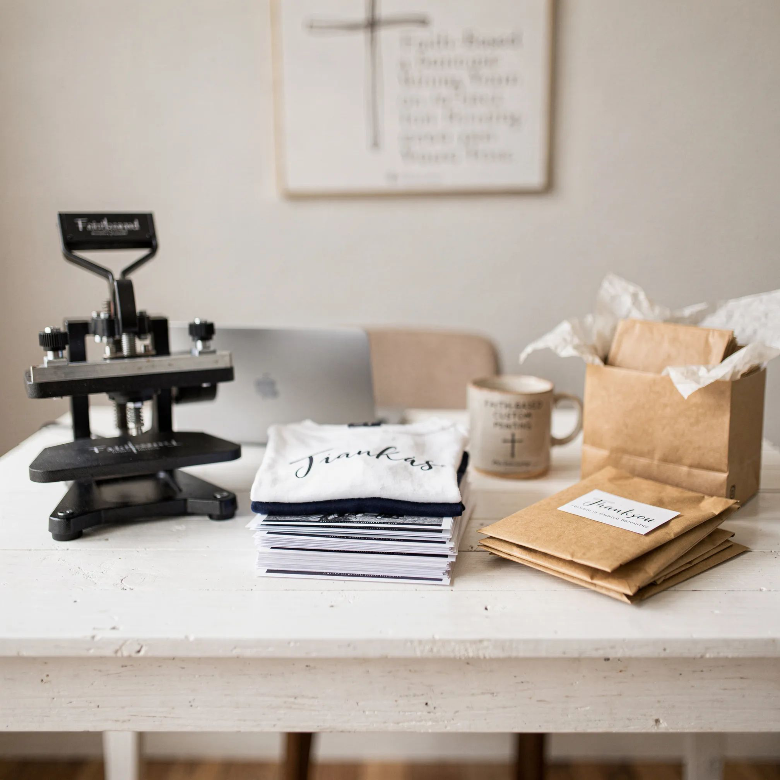 A white wooden table with a black pressing iron, a stack of thank you cards, a paper bag filled with tissue paper, a coffee mug with religious text, a laptop, and a box with tissue paper, in a room with wall decor.
