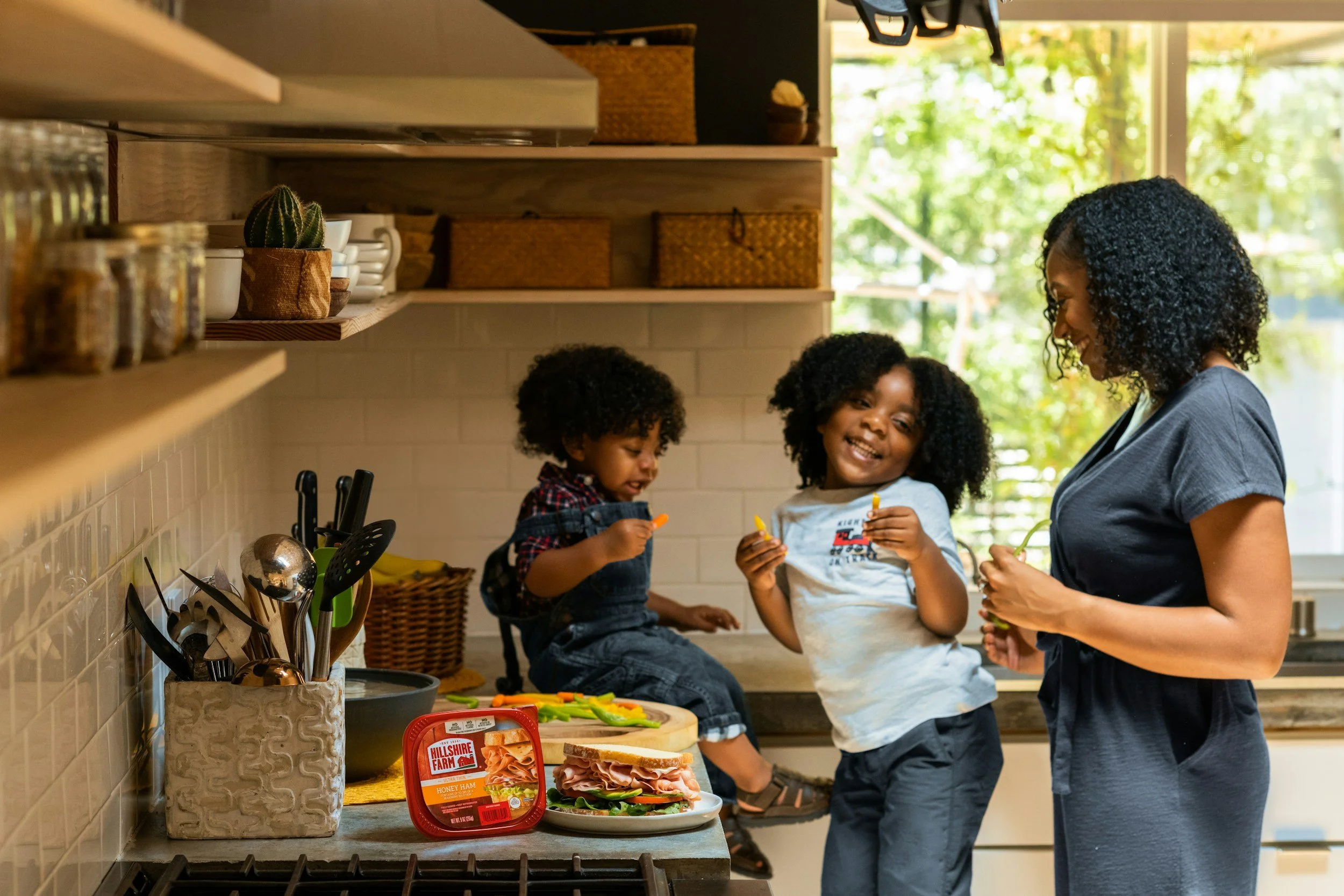 A woman with two children in a kitchen, preparing sandwiches with various ingredients on the counter. The children are holding slices of cheese, smiling, and sitting on the kitchen counter, while the woman stands nearby. The kitchen has open shelves with jars and baskets, and sunlight streams through a window with green trees outside.