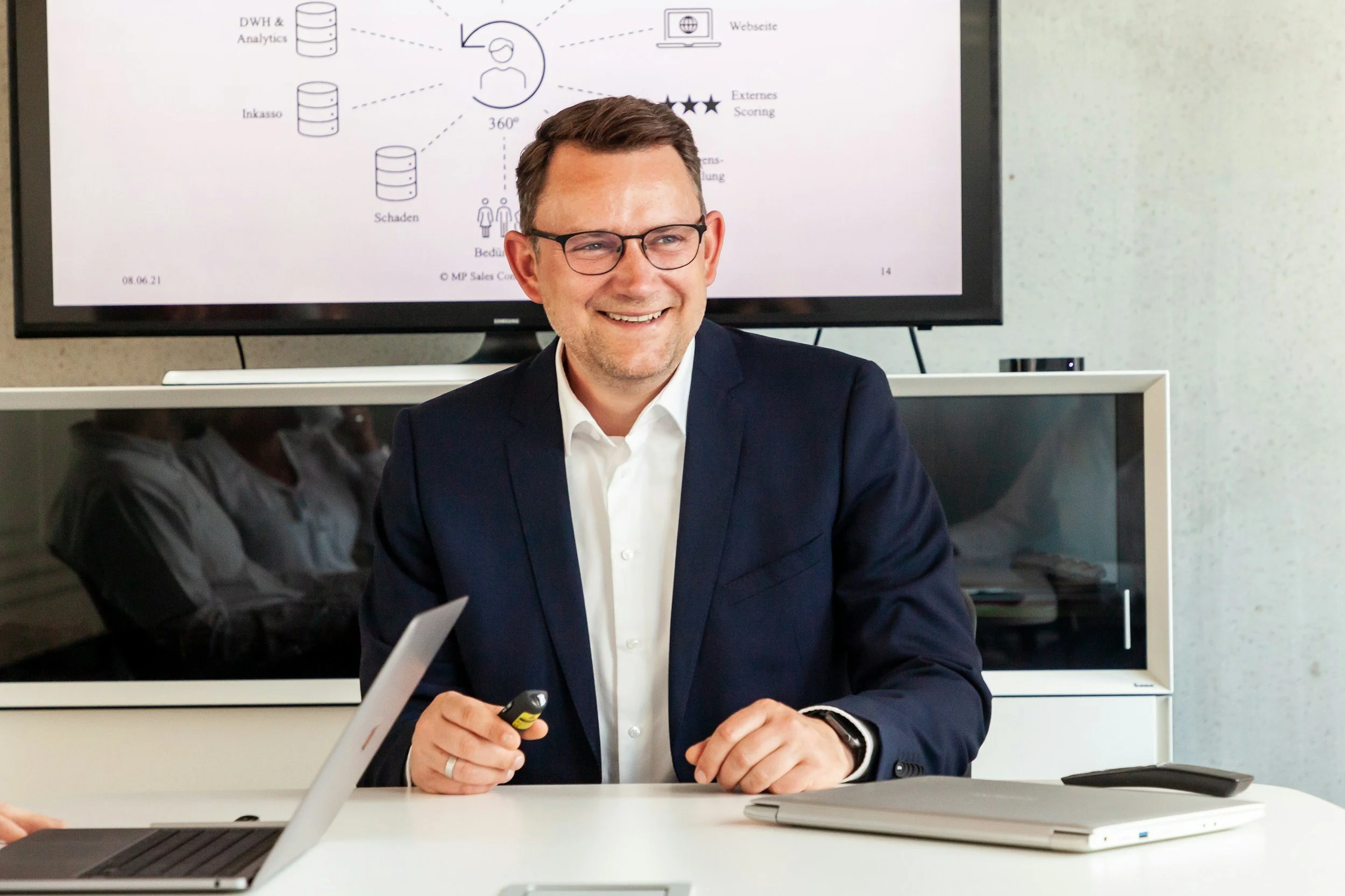A smiling man in a dark suit and glasses sitting at a table with a laptop, a notebook, and a remote, in a conference room with a large screen displaying a presentation