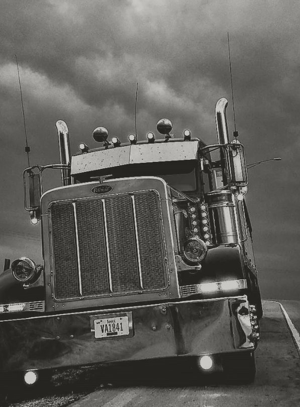 Black and white photo of a large semi-truck with a prominent front grille, multiple lights, and multiple antennas, facing the camera on a road under a cloudy sky.