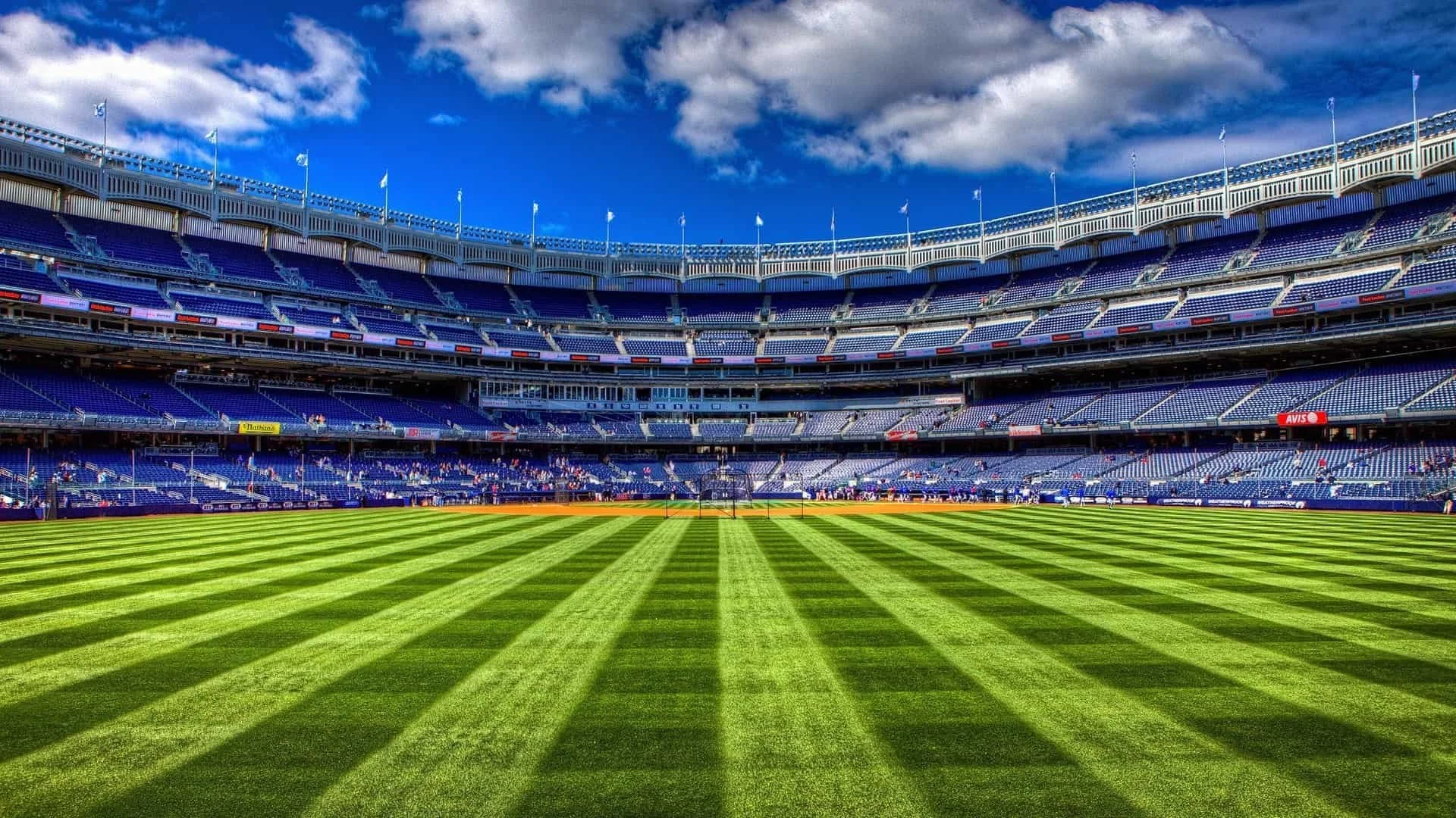 Empty baseball stadium with a green field, multi-level blue seats, and a partly cloudy sky overhead.